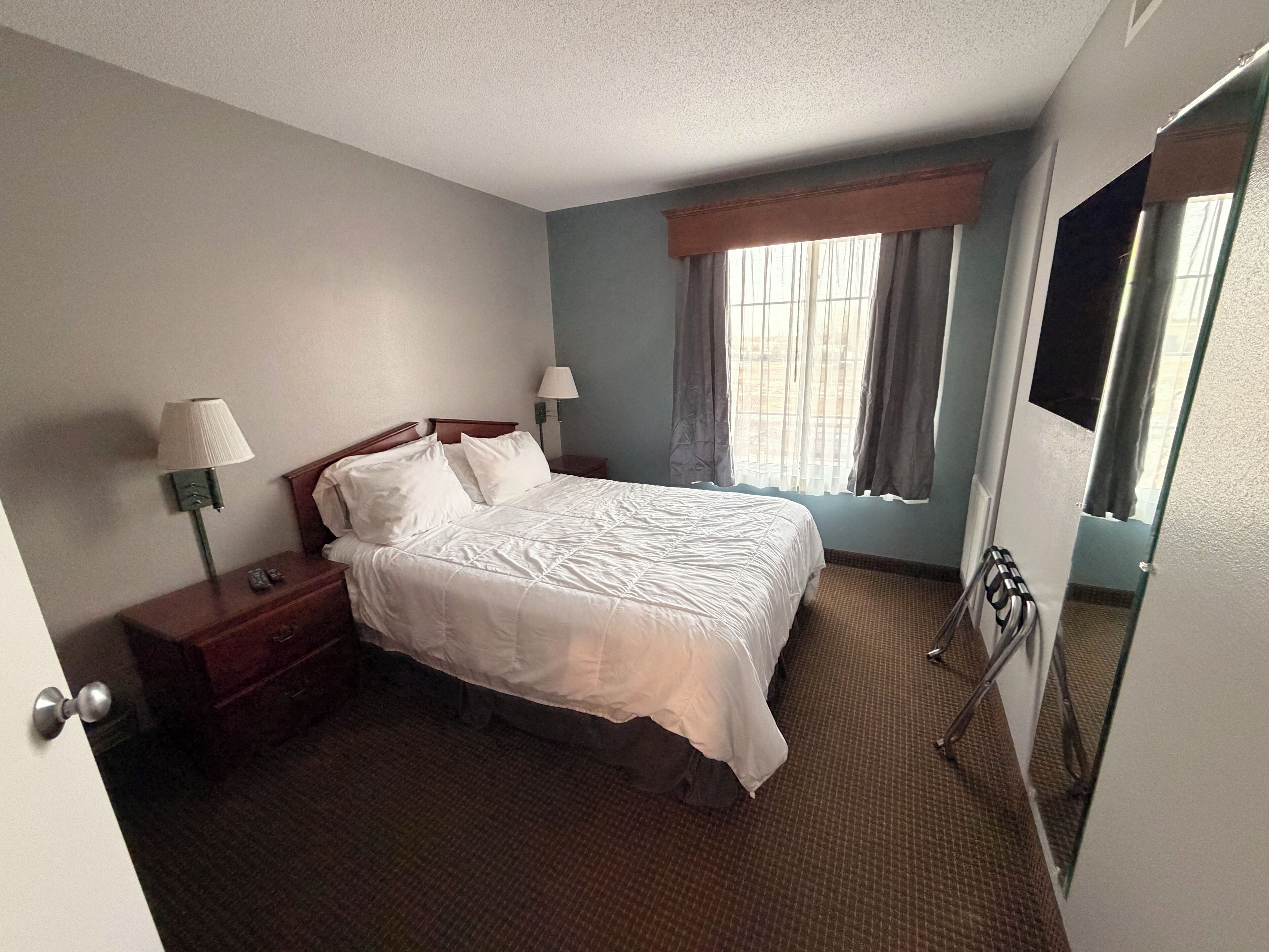 A hotel room with a large bed, two bedside tables with lamps, a window with curtains, and a door with a mirror.