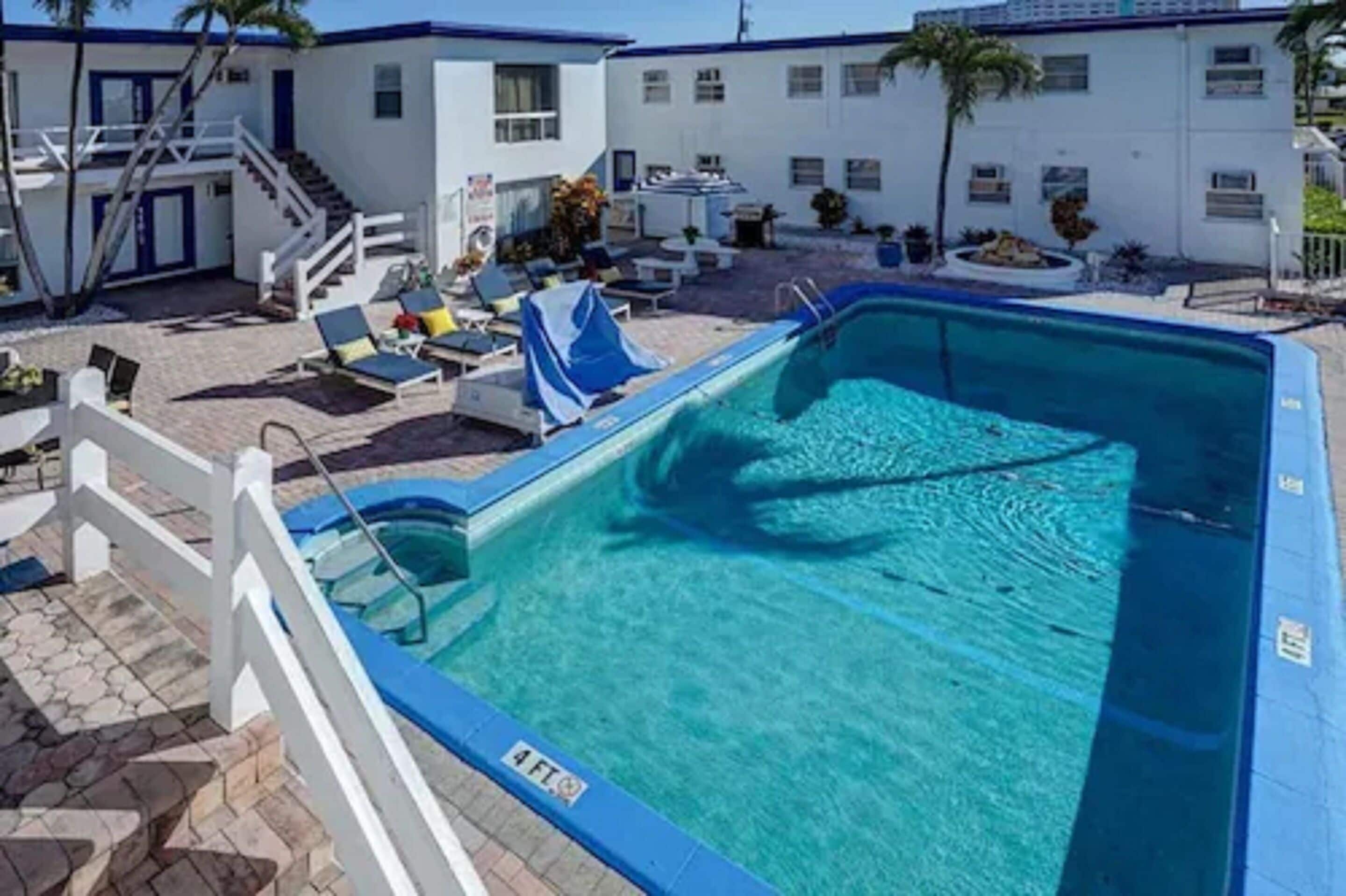 A swimming pool with lounge chairs and a covered area, surrounded by a white building with a blue roof and a staircase leading to an upper level.