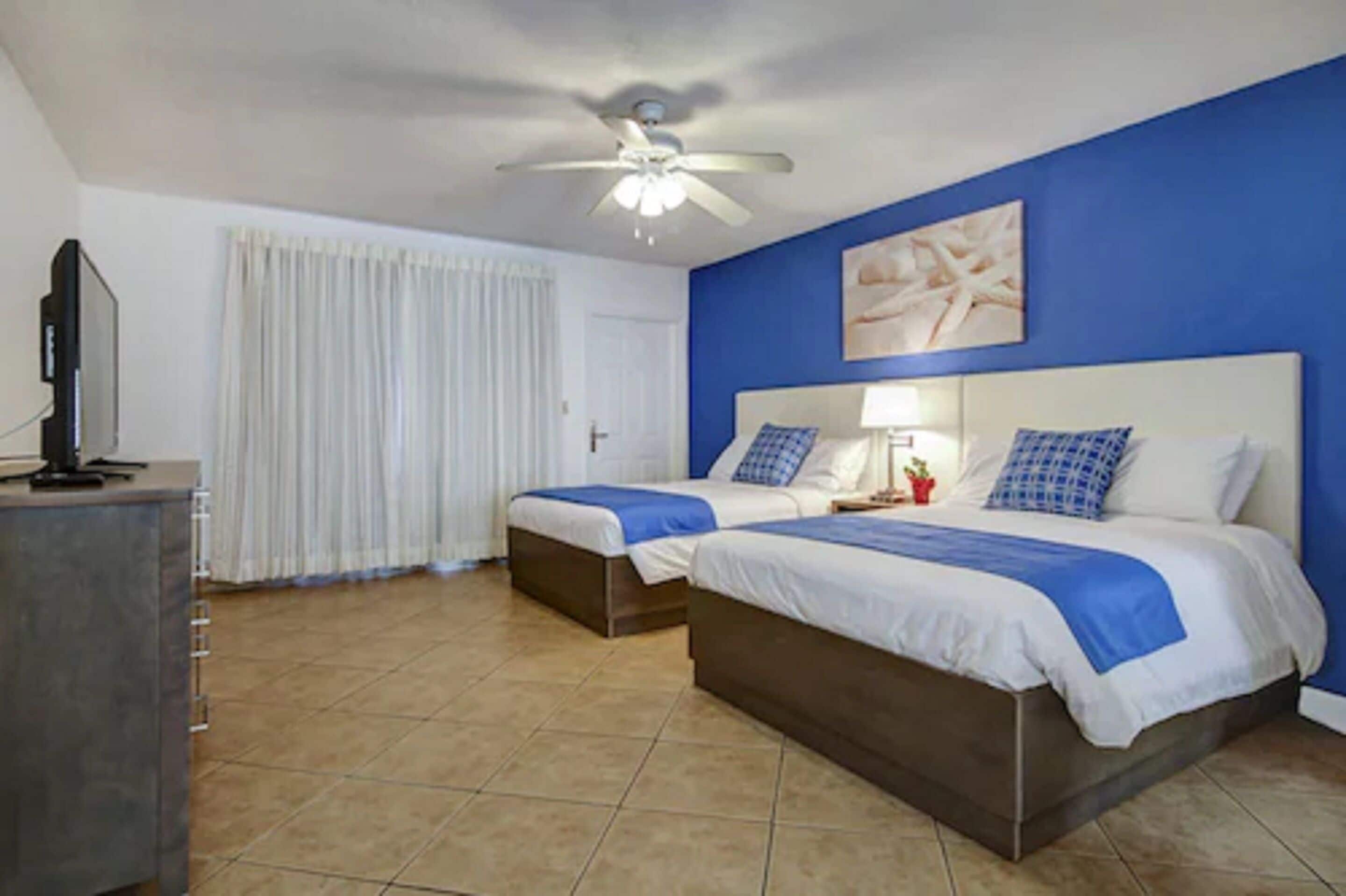 A hotel room with two beds, a ceiling fan, a television, and a window with curtains.