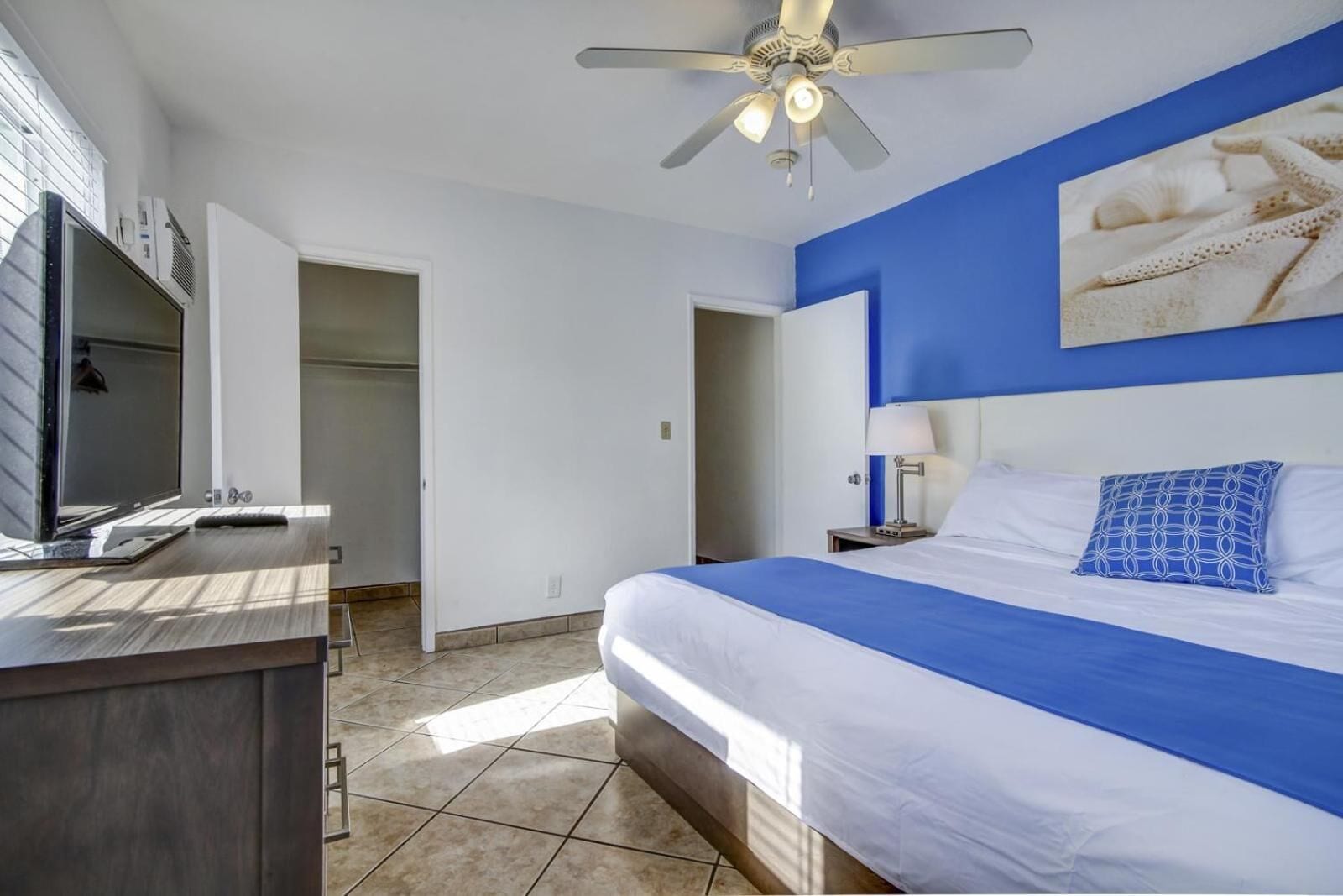 A hotel room with a bed, a desk with a computer, a ceiling fan, and a large window.