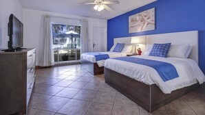 A hotel room with two beds, a TV, a ceiling fan, a large window with curtains, and a blue wall.