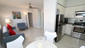 A modern living room with a grey sofa, a red pillow, and a glass table with a candle. A kitchen with stainless steel appliances is visible in the background.