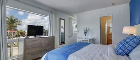 A hotel room with a bed, a TV on a wooden dresser, a desk with a lamp, and a view of palm trees through large windows.