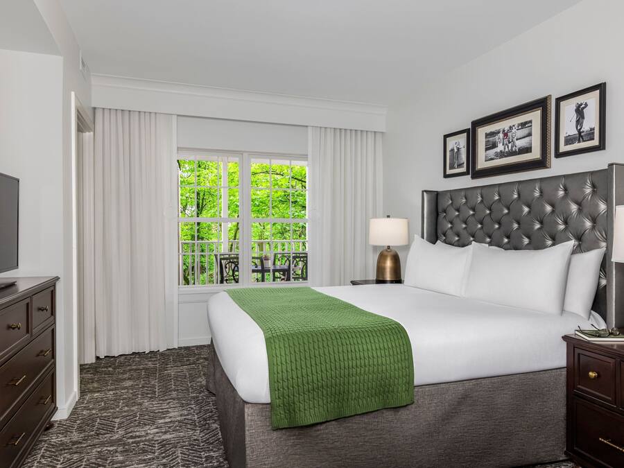 A hotel room with a large bed, a television, a nightstand with lamps, and a window with curtains.