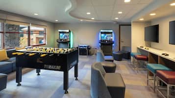 A room with a foosball table, two arcade game machines, a flat-screen TV, and a bar area with stools.