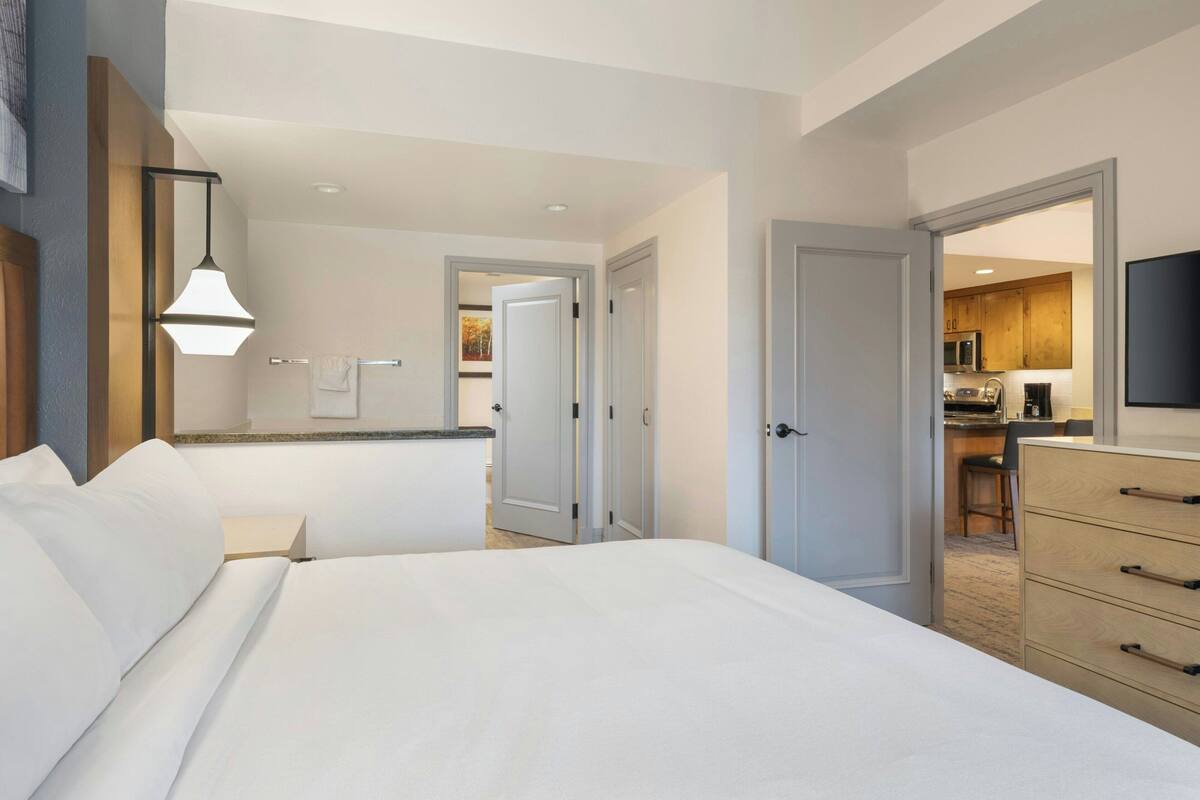 A hotel room with a large bed, a desk with a chair, a flat-screen TV, and a kitchenette in the background.