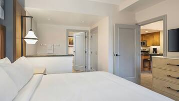 A hotel room with a large bed, a desk with a chair, a flat-screen TV, and a kitchenette in the background.