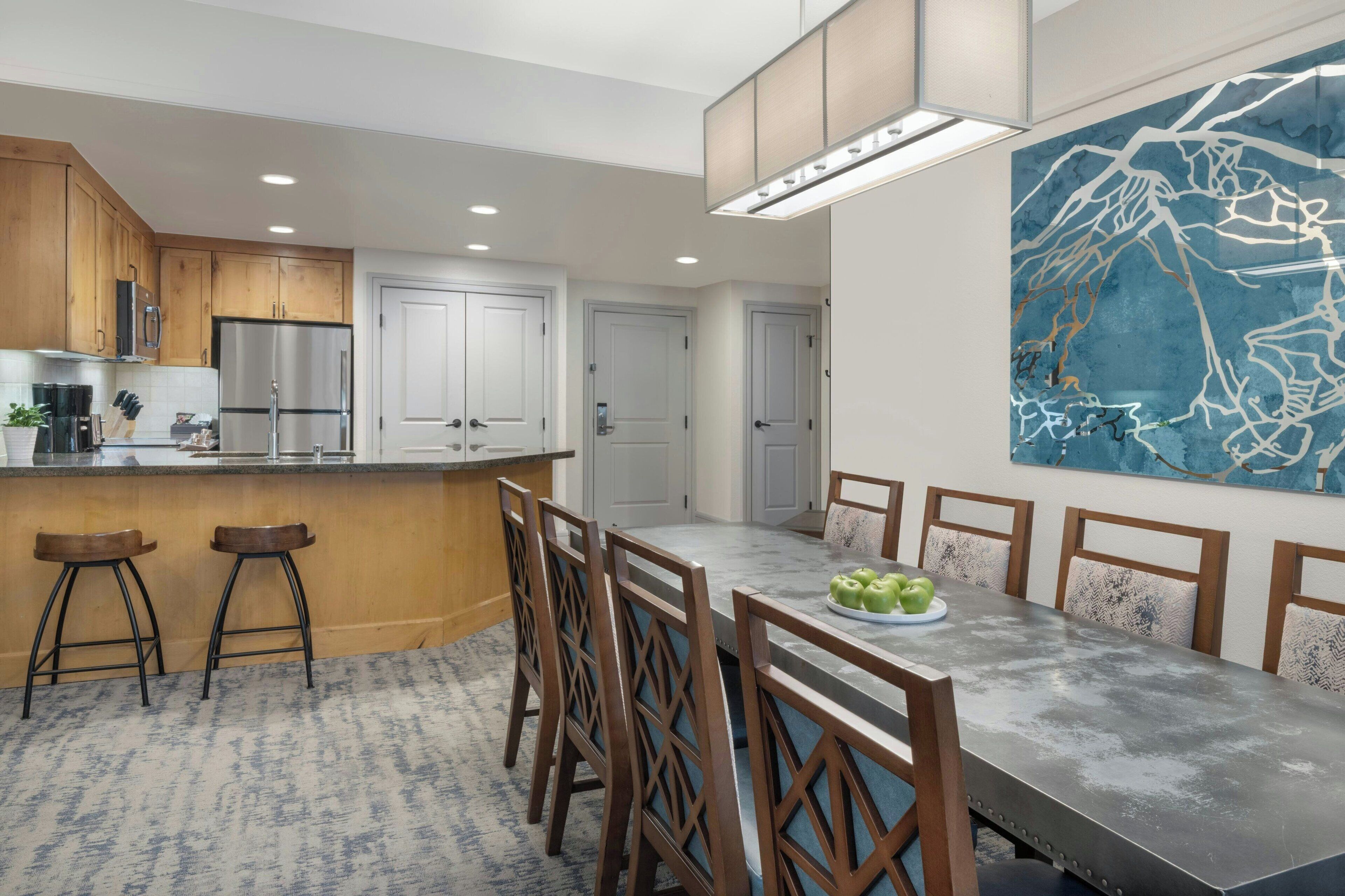 A modern kitchen with wooden cabinets, a stainless steel refrigerator, and a dining area with a long table and chairs.