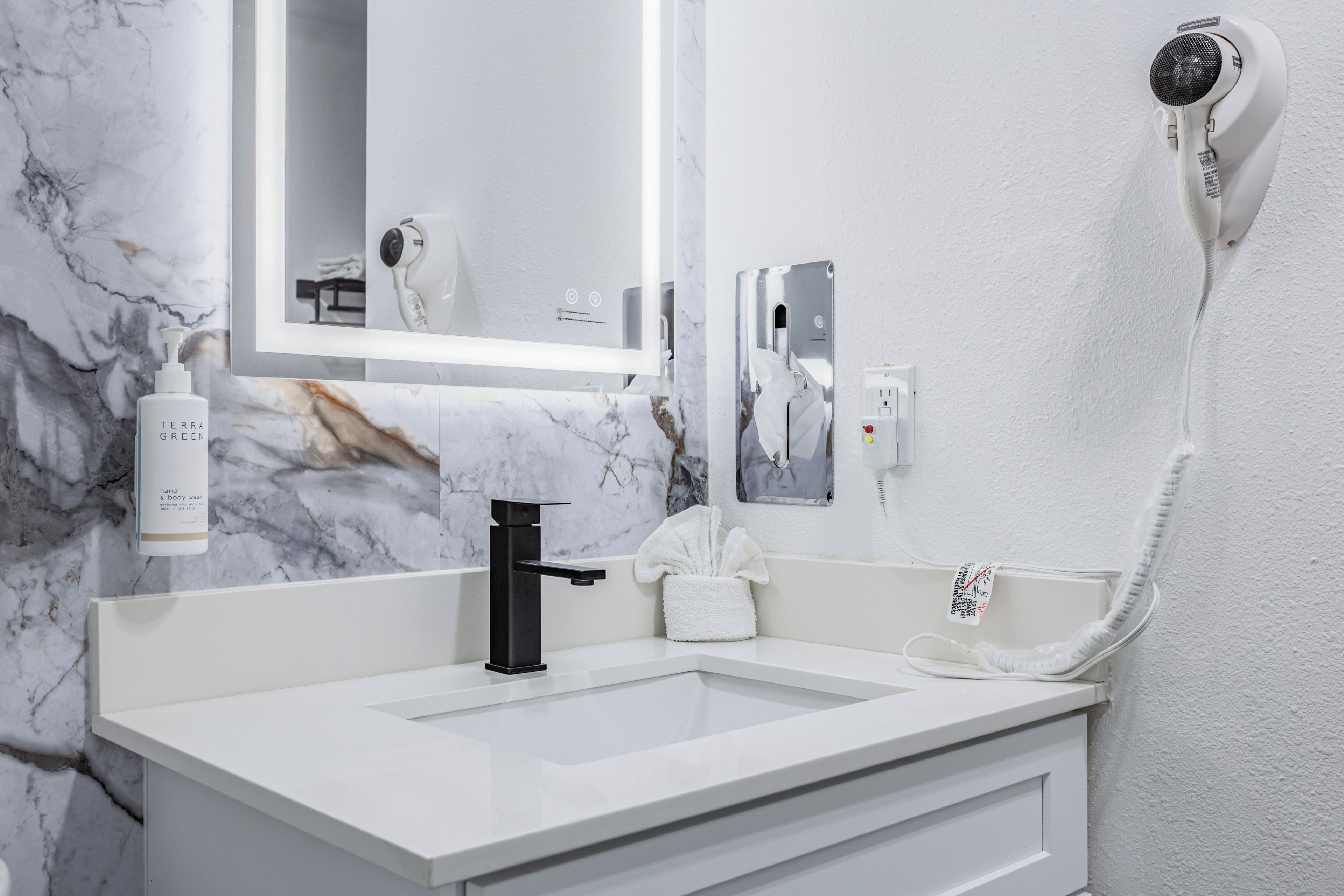 Une salle de bain avec un lavabo blanc, un robinet noir, un sèche-mains, un porte-serviettes et un flacon de savon liquide.