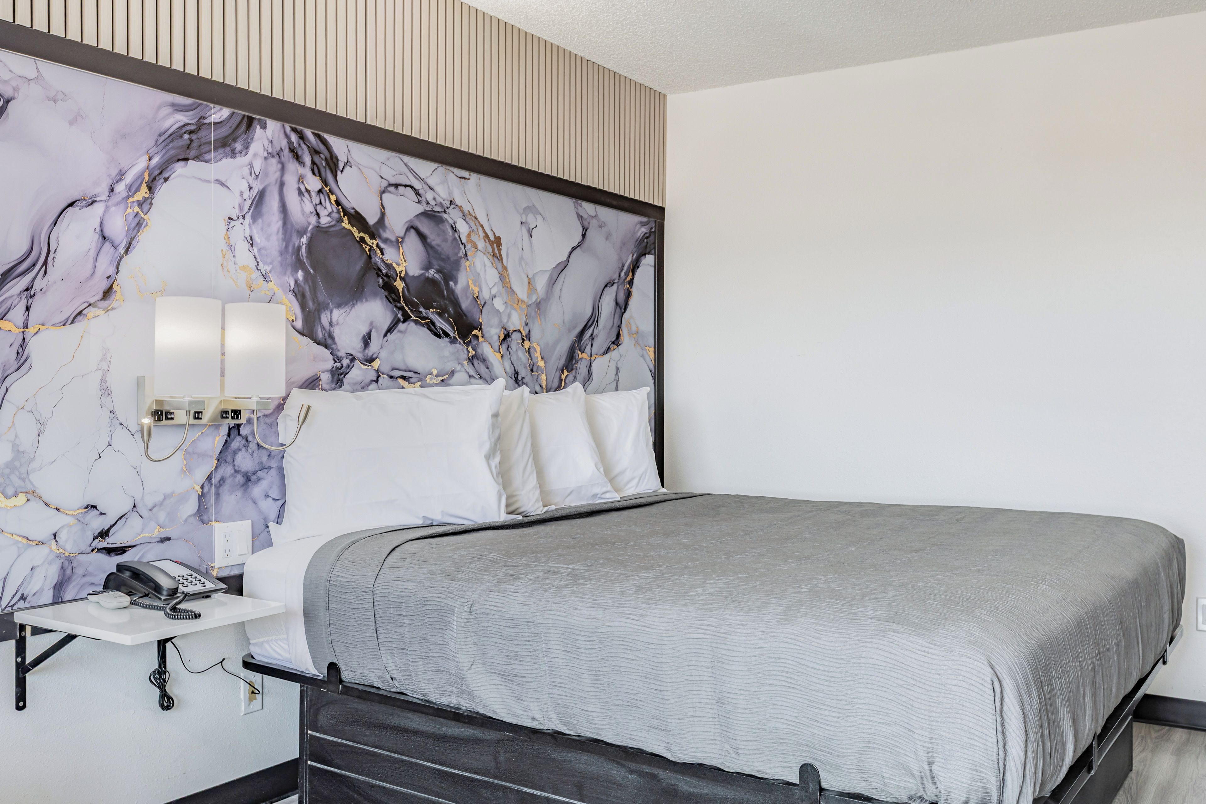 Una habitación de hotel moderna con cabecera de diseño de mármol, mesita de noche con lámpara y colcha gris.