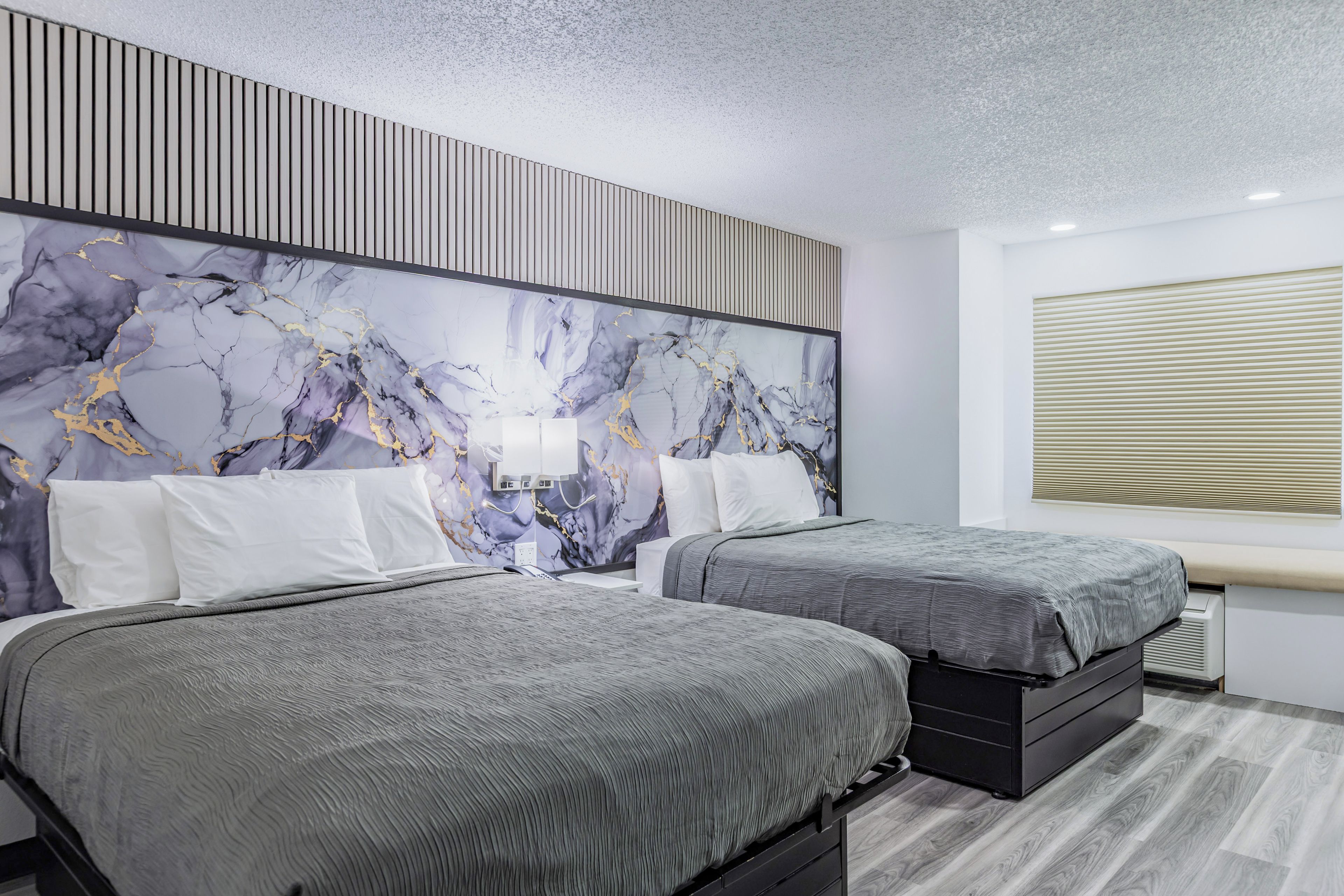 A modern hotel room with two beds, a marble-patterned headboard, a large window with blinds, and a built-in heater.