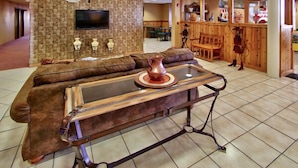 A lobby with a brown leather sofa, a wooden coffee table with a copper vase, and a television mounted on a textured wall. - Western Skies Inn And Suites (Los Lunas)