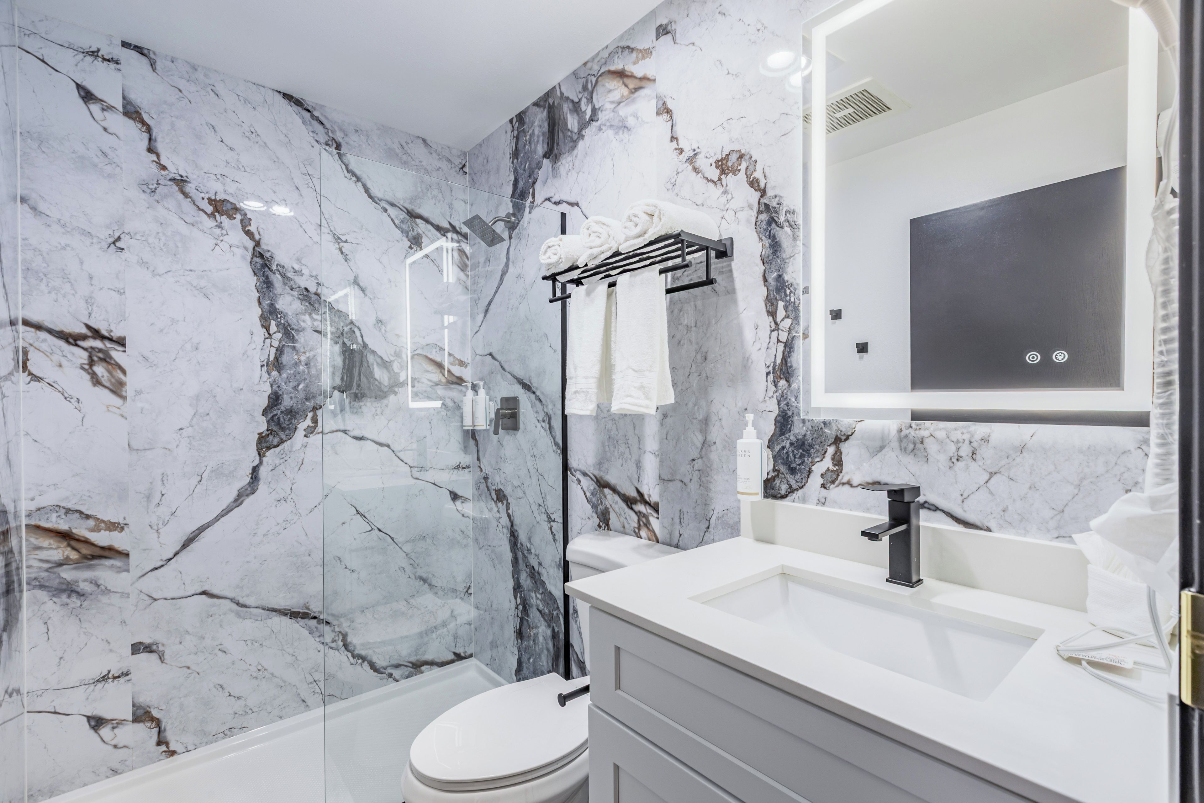 A modern bathroom with a marble wall, a glass-enclosed shower, a white toilet, and a vanity with a mirror.