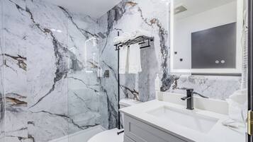A modern bathroom with a marble wall, a glass-enclosed shower, a white toilet, and a vanity with a mirror.