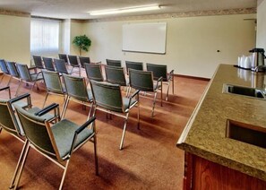 A conference room with rows of chairs, a whiteboard, and a kitchenette area. - Western Skies Inn And Suites (Los Lunas)