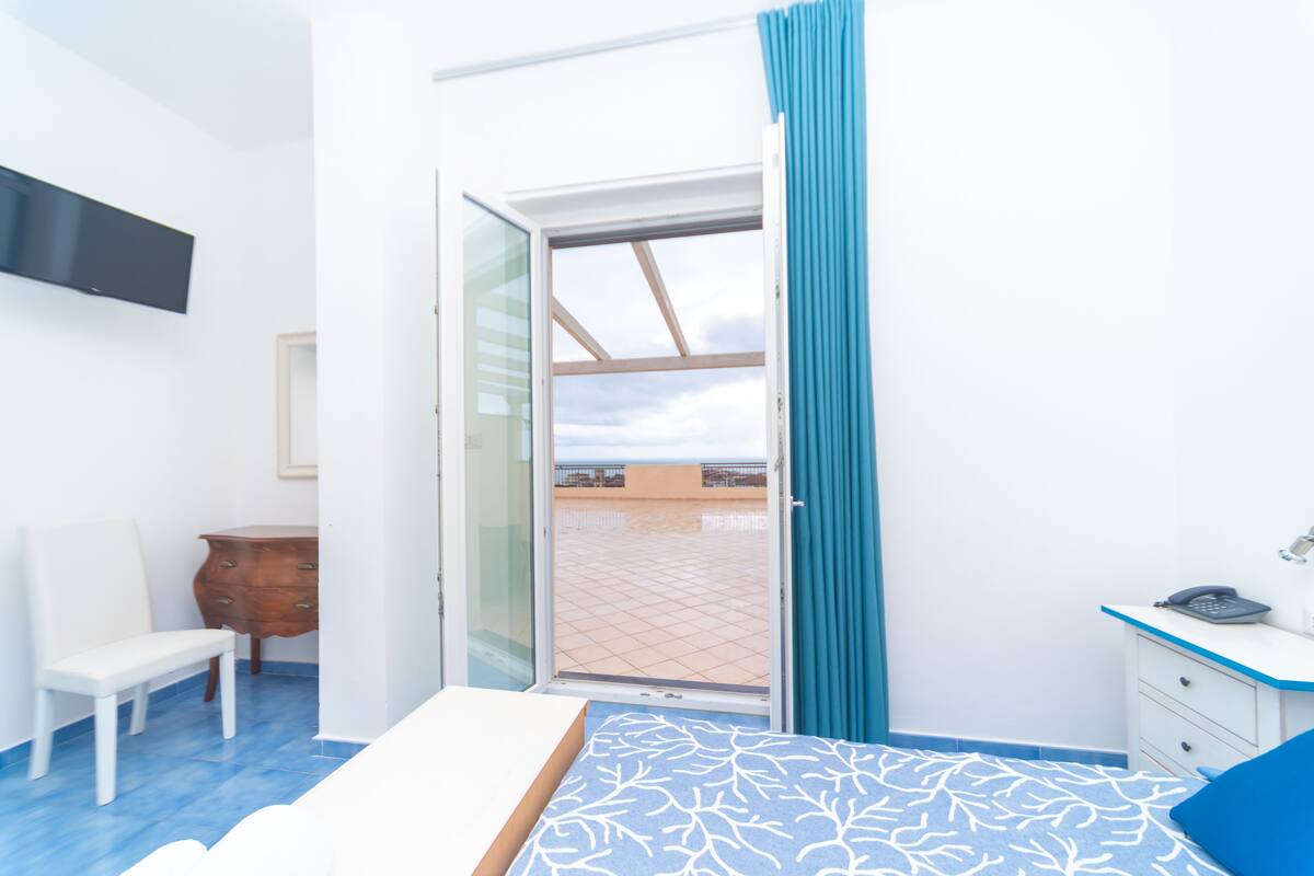 A room with a bed, a desk, a chair, a television, and a sliding glass door to an outdoor area.