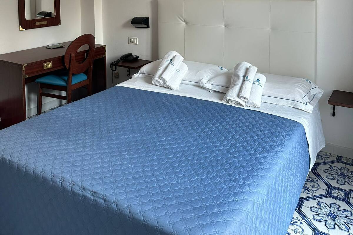 A hotel room with a bed, a desk, a chair, a mirror, a telephone, and a painting on the wall.