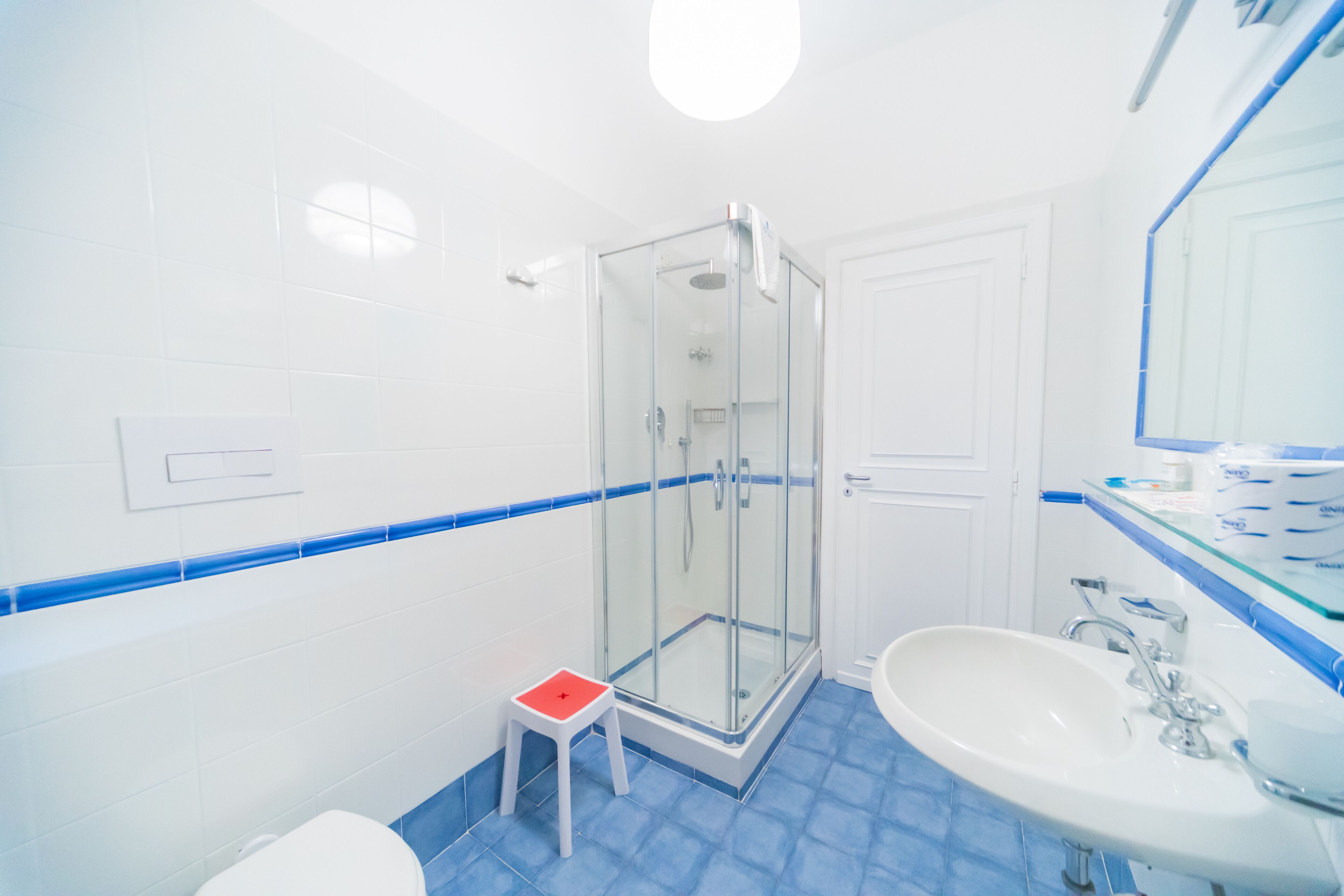 A bathroom with a glass shower enclosure, a white sink, and a small red stool.