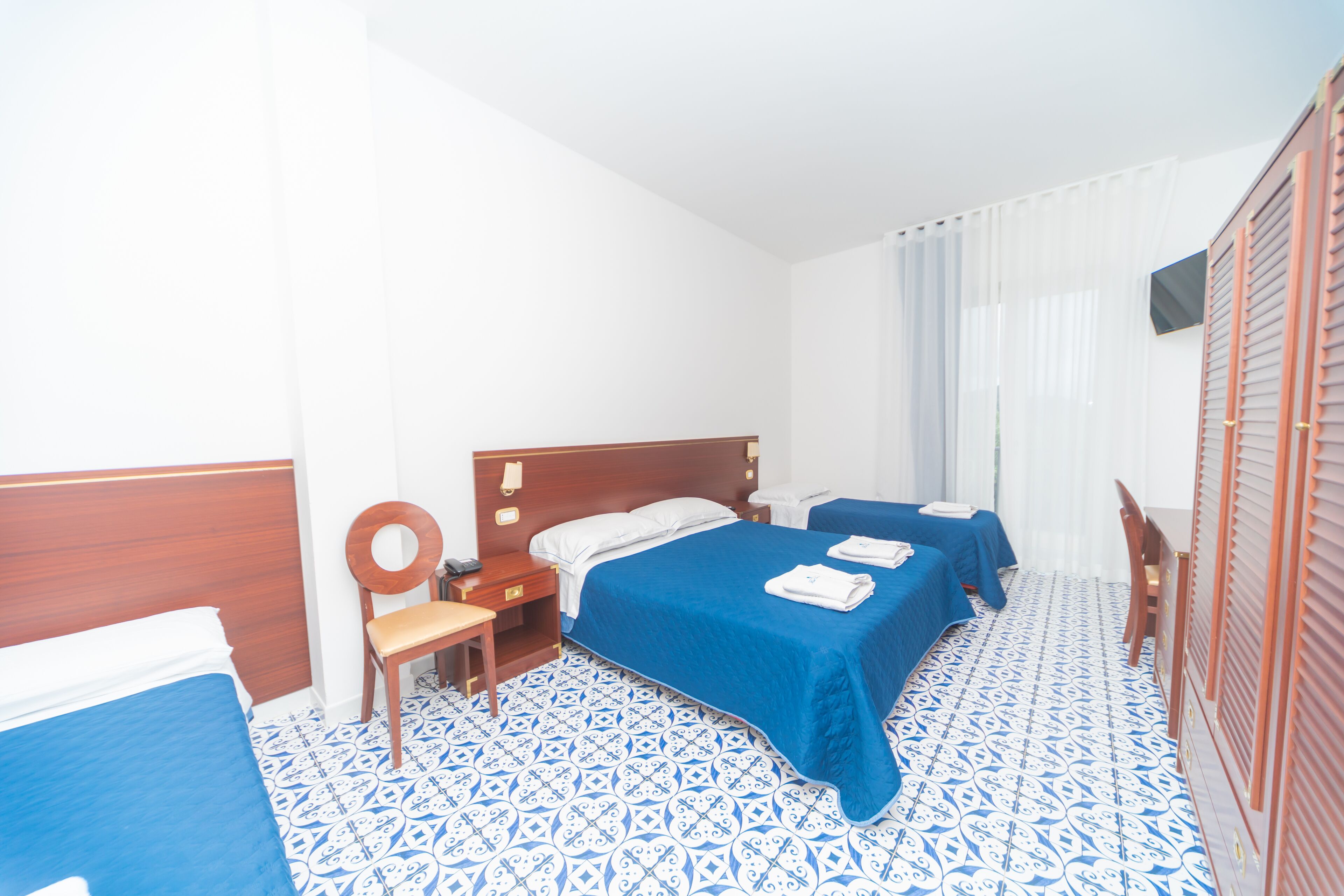 A hotel room with two beds, a wooden headboard, a chair, a small table, and a window with curtains.