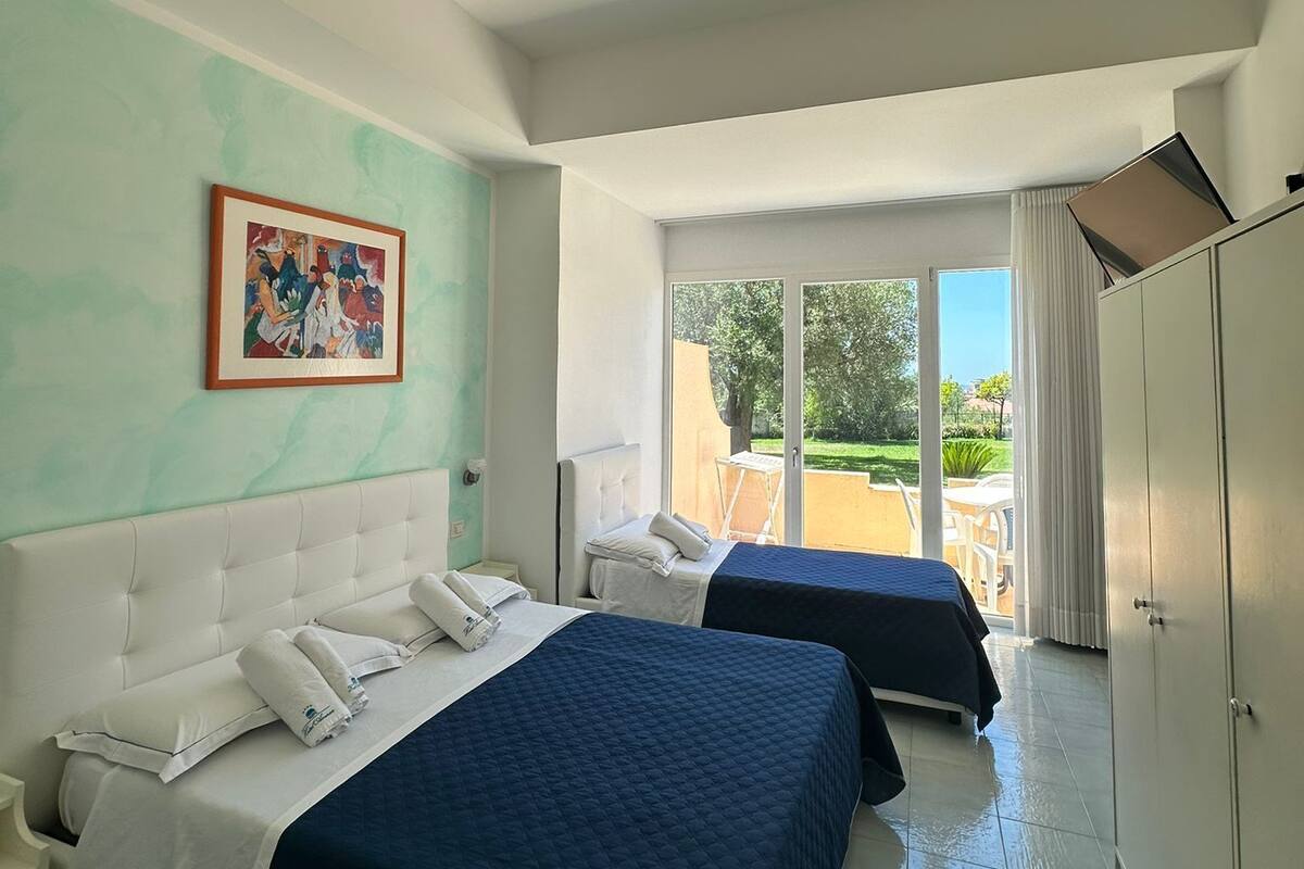 A hotel room with two beds, a painting on the wall, a ceiling fan, and a view of the outdoors.