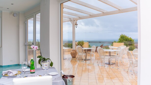 A balcony with a table set for dining, chairs, and a view of the sea. - Hotel America (Camerota)