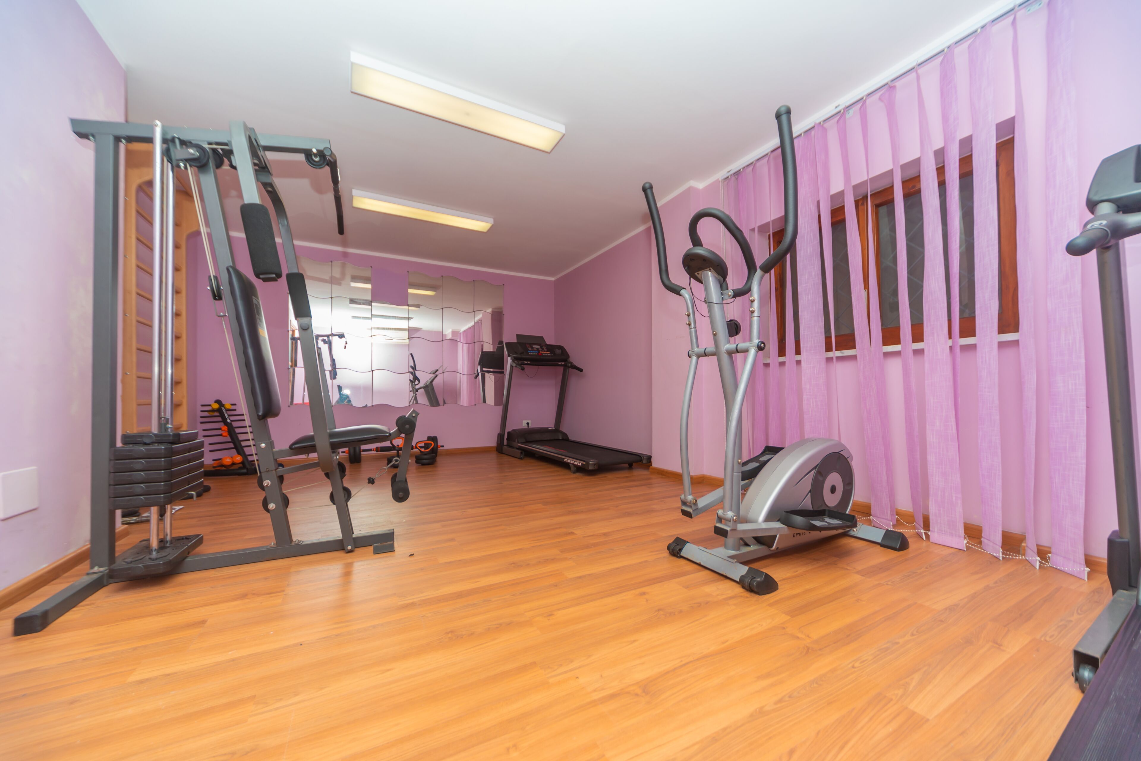 Un gimnasio con equipo de ejercicio, piso de madera y cortinas rosas.
