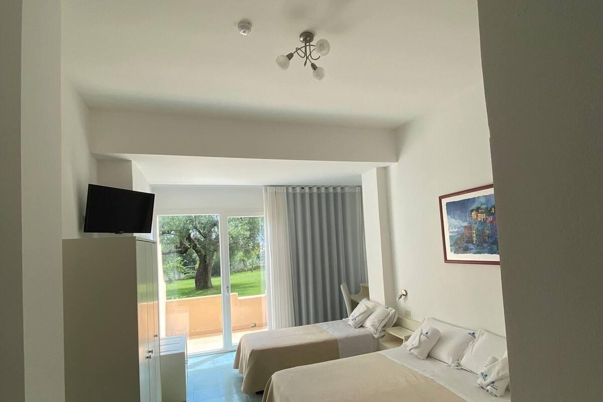A hotel room with two beds, a television on a cabinet, a ceiling fan, and a view of the outdoors.