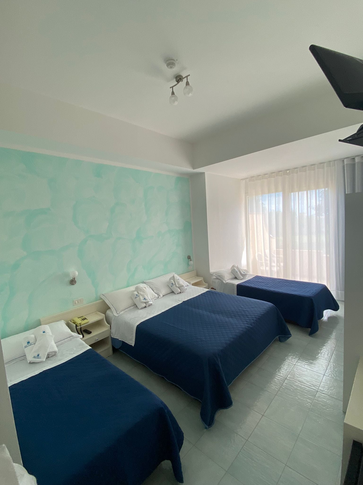 A hotel room with three beds, a window with sheer curtains, and a ceiling light fixture.