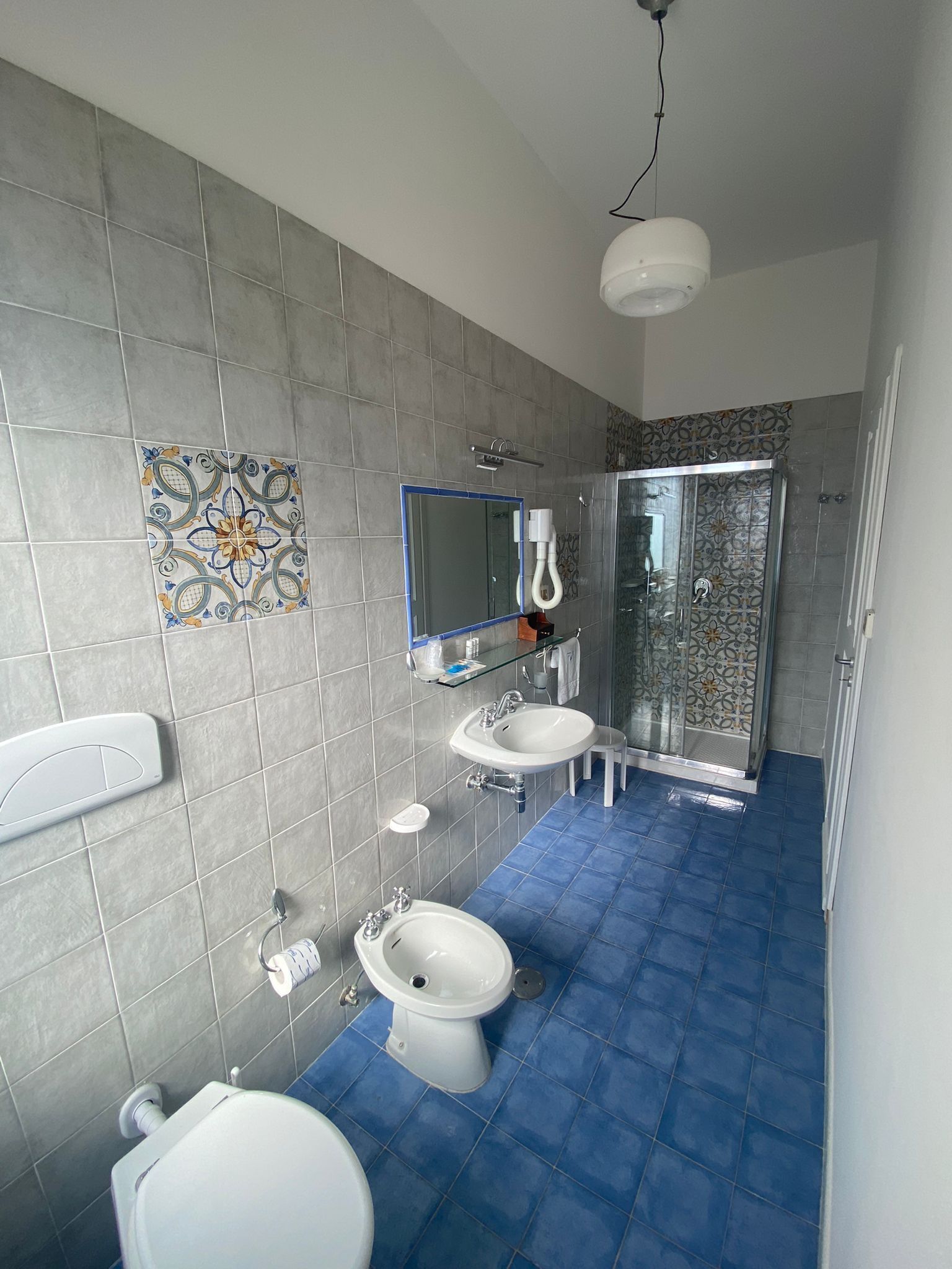 A bathroom with grey tiled walls, a blue-tiled floor, a white toilet, two sinks, a shower enclosure, and a decorative tile on the wall.