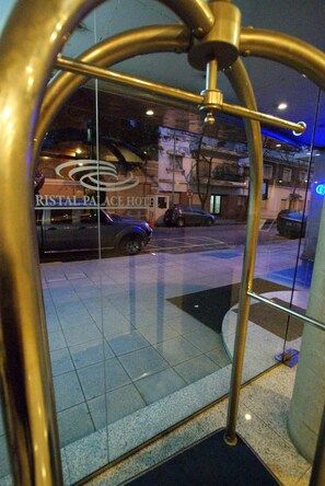 Entrada con puerta de cristal y marco metálico que refleja una vista de la calle y un edificio con el letrero que dice "RISTAL PALACE HOTEL".