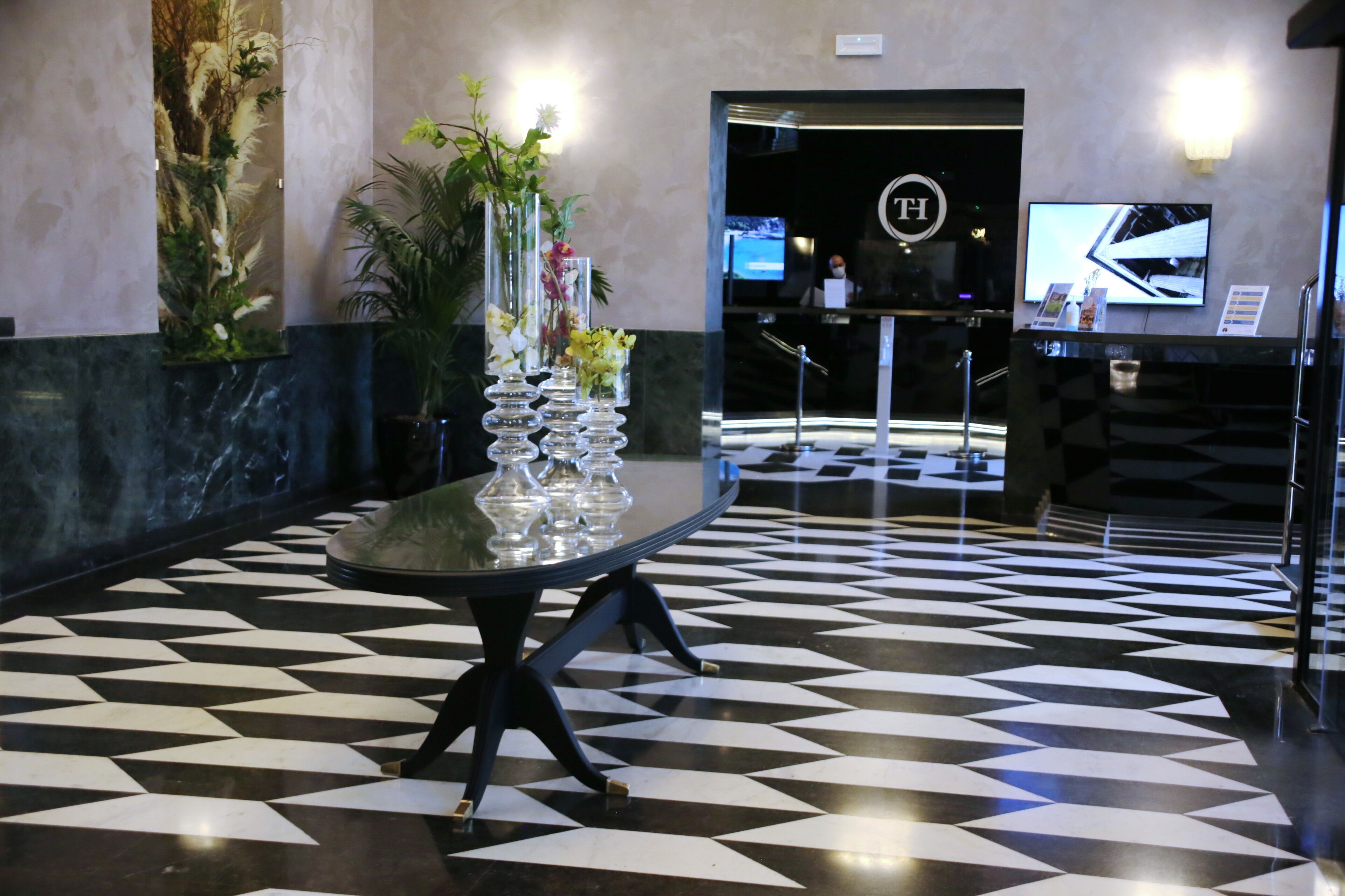 A lobby with a checkered floor, a reception desk, and a large plant with flowers.
