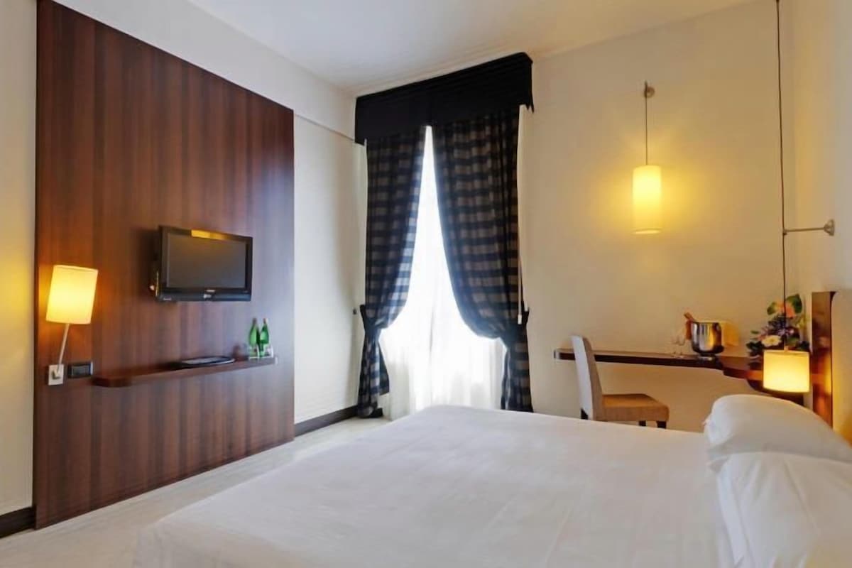 Habitación de hotel con una cama grande, un televisor de pantalla plana montado en la pared, un escritorio con una silla y una ventana con cortinas.
