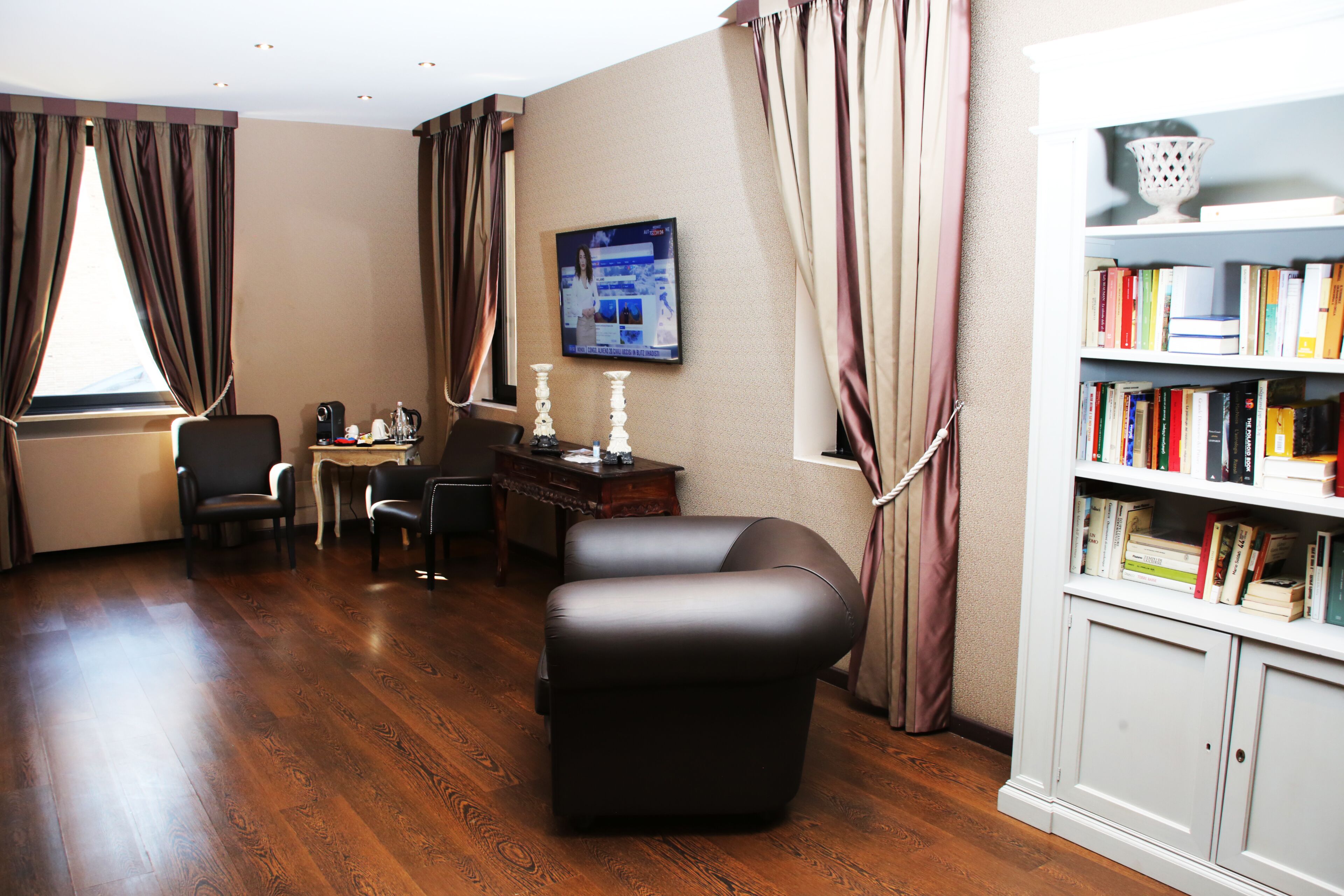 A hotel room with a wooden floor, a leather armchair, a desk with a computer, a bookshelf, and a large window with curtains.