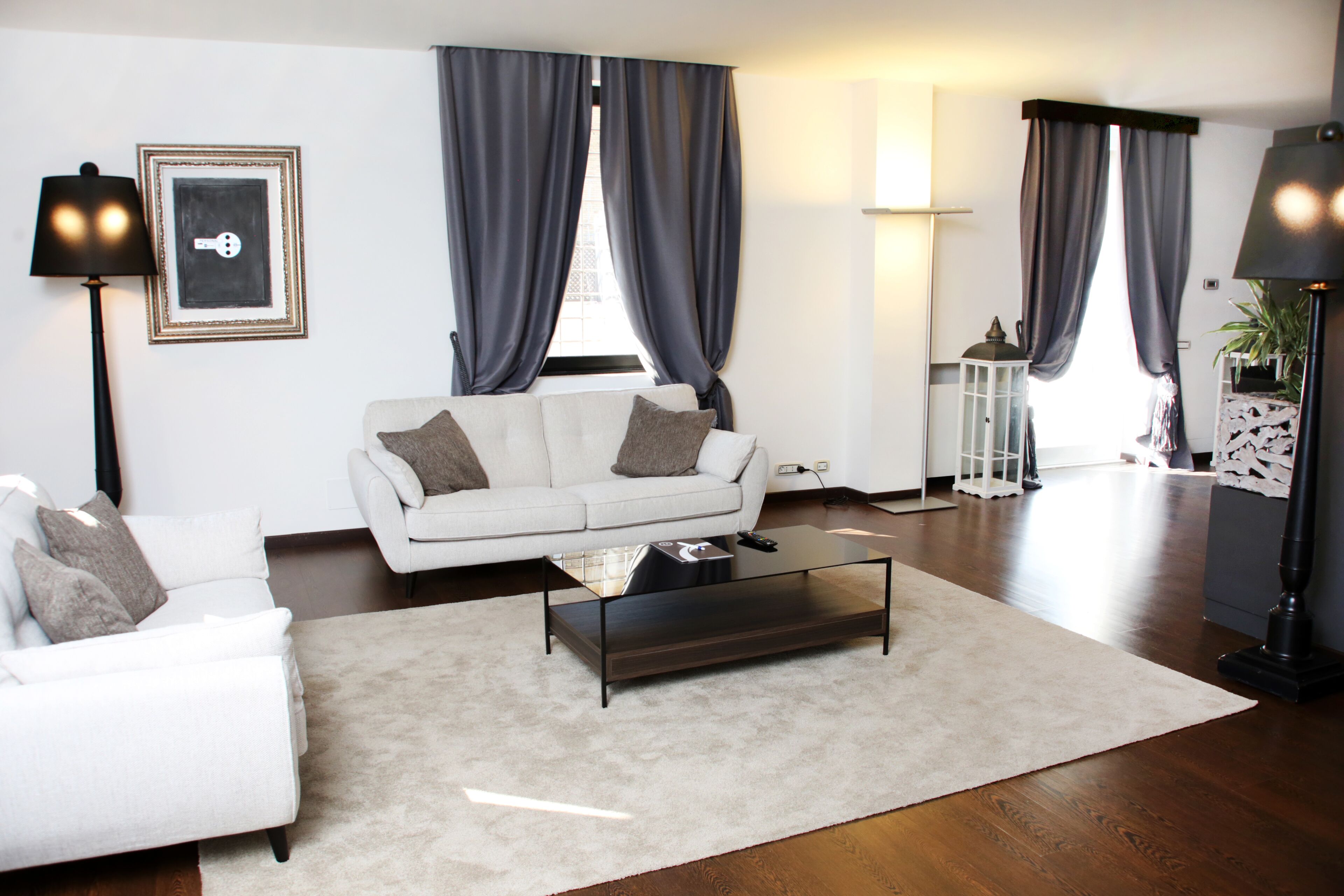 A modern living room with a white sofa, a glass coffee table, and a large window with dark curtains.