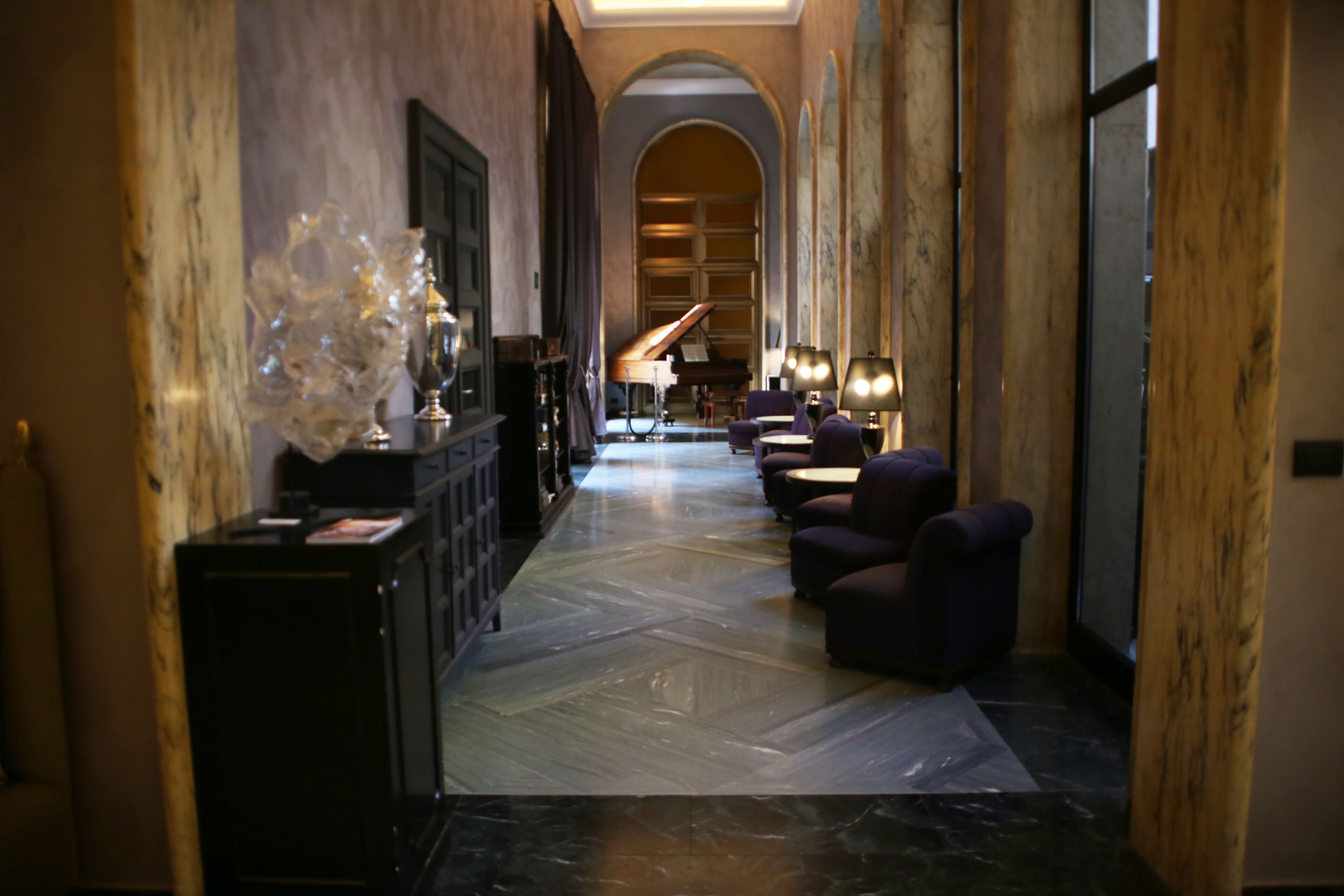 A hallway with a grand piano, plush seating, and a marble floor.
