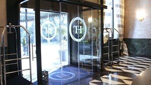 A hotel entrance with a glass door, a metal cart, and a patterned floor. - TH Roma - Carpegna Palace (Rome)