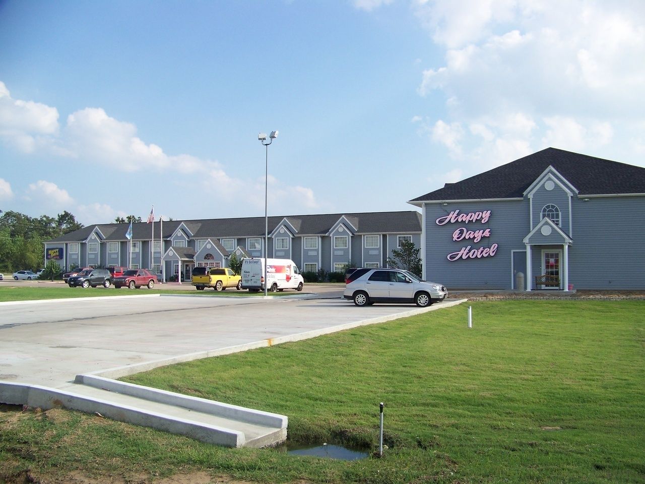 A hotel with a sign reading "Happy Days Hotel" and a parking lot with several vehicles.