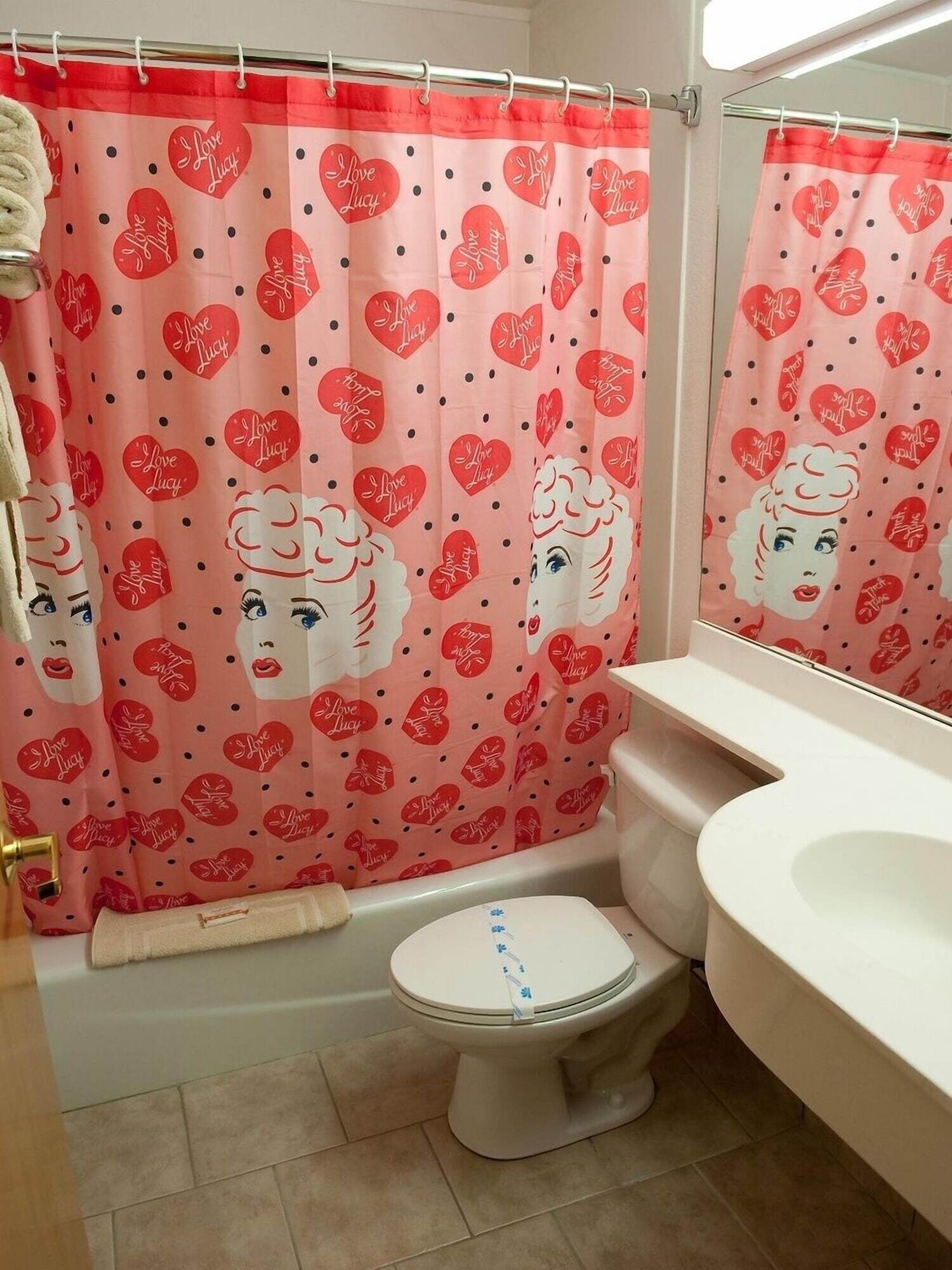 Une salle de bain avec un rideau de douche rose orné de cœurs et d’un motif représentant un visage, des toilettes blanches et un lavabo blanc.