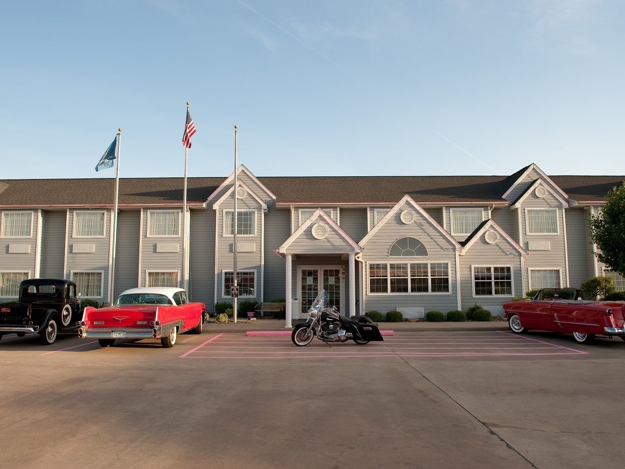 A motel with a central entrance, a parking lot with vintage cars, and a flagpole with flags.