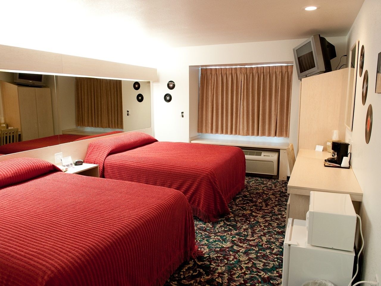 Ein Hotelzimmer mit zwei Betten, einem Schreibtisch, einem Spiegel, einem Fernseher und einem Fenster mit Vorhängen.