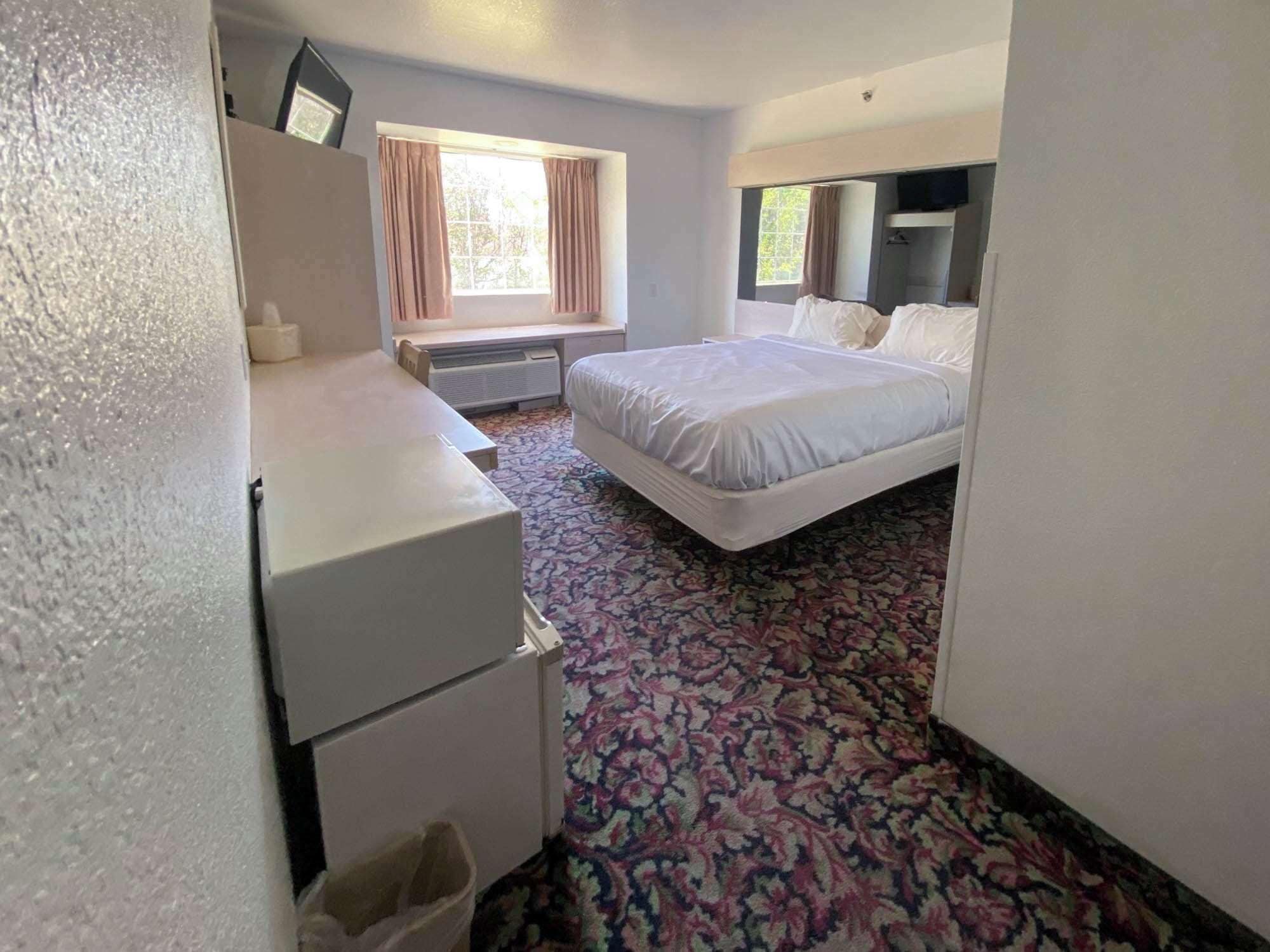 A hotel room with a bed, a desk, a chair, a small refrigerator, and a window with curtains.