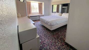 A hotel room with a bed, a desk, a chair, a small refrigerator, and a window with curtains.