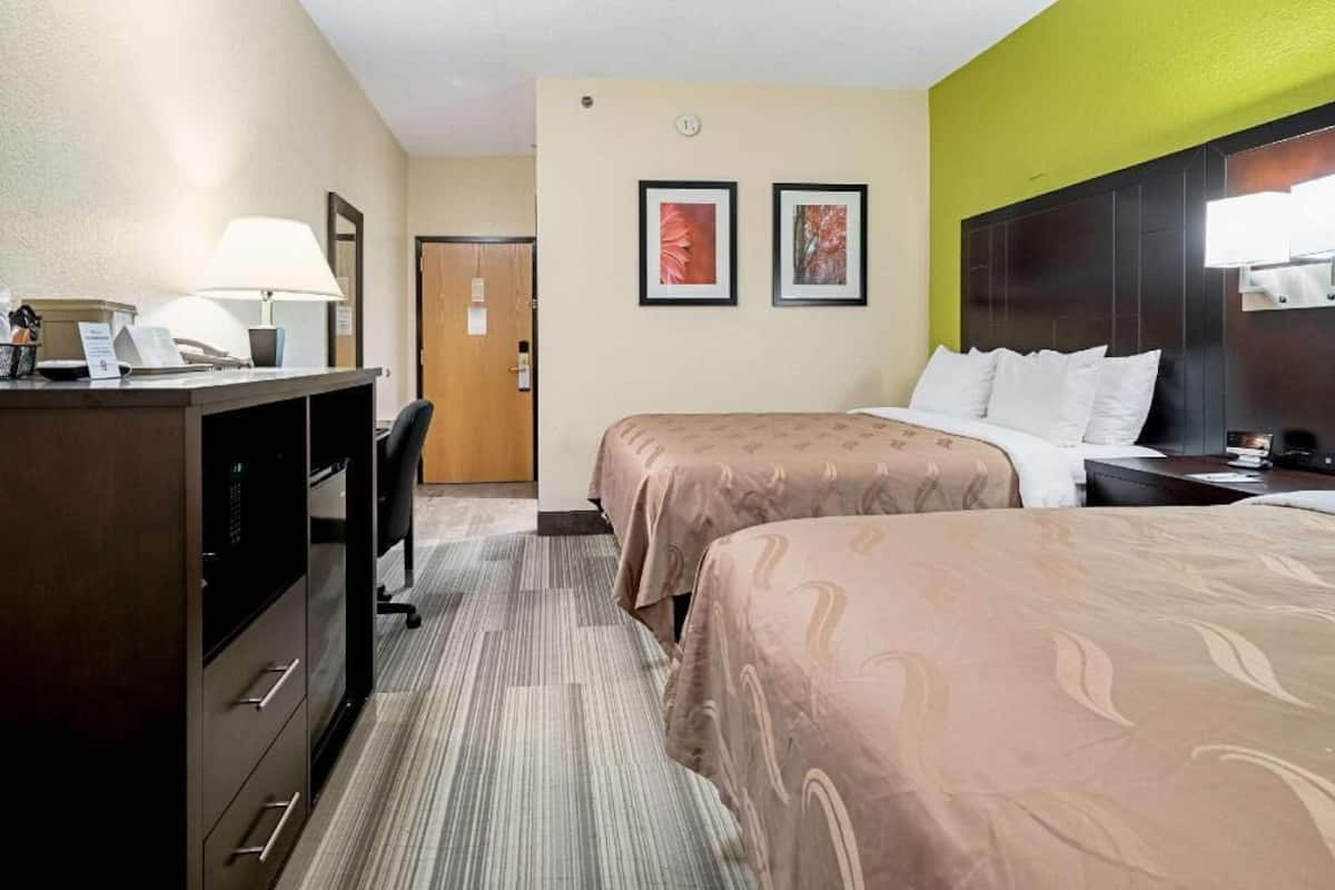 A hotel room with two beds, a desk with a lamp, a chair, and a door.