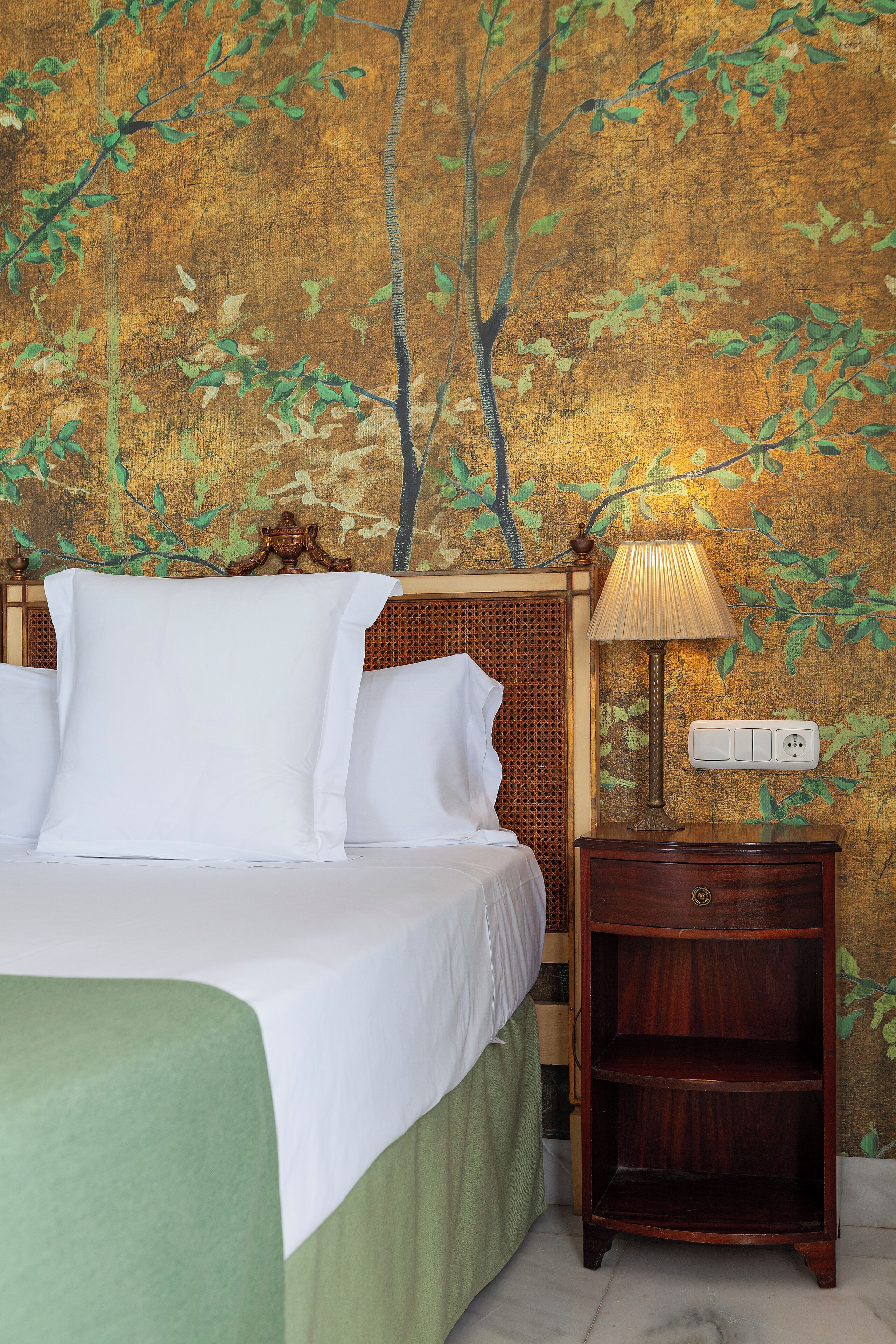 A hotel room with a bed, a nightstand with a lamp, and a wall with a tree mural.