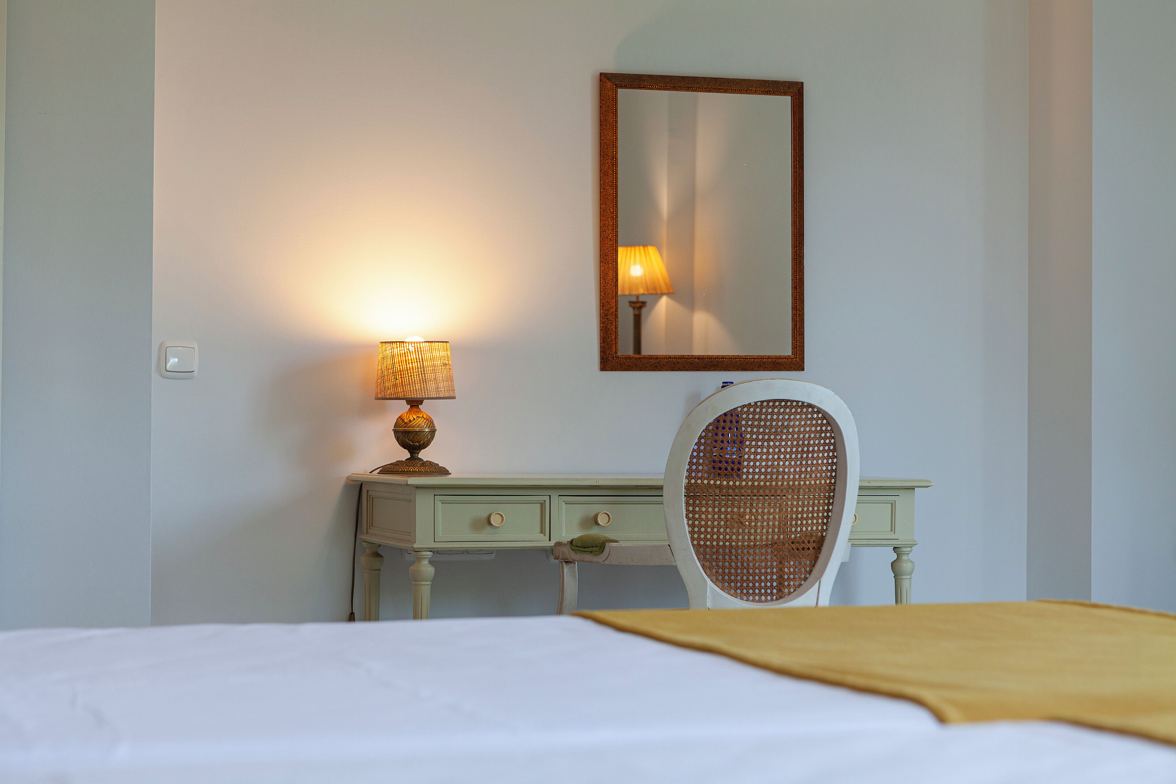 A hotel room with a bed, a desk with drawers, a lamp, and a mirror.