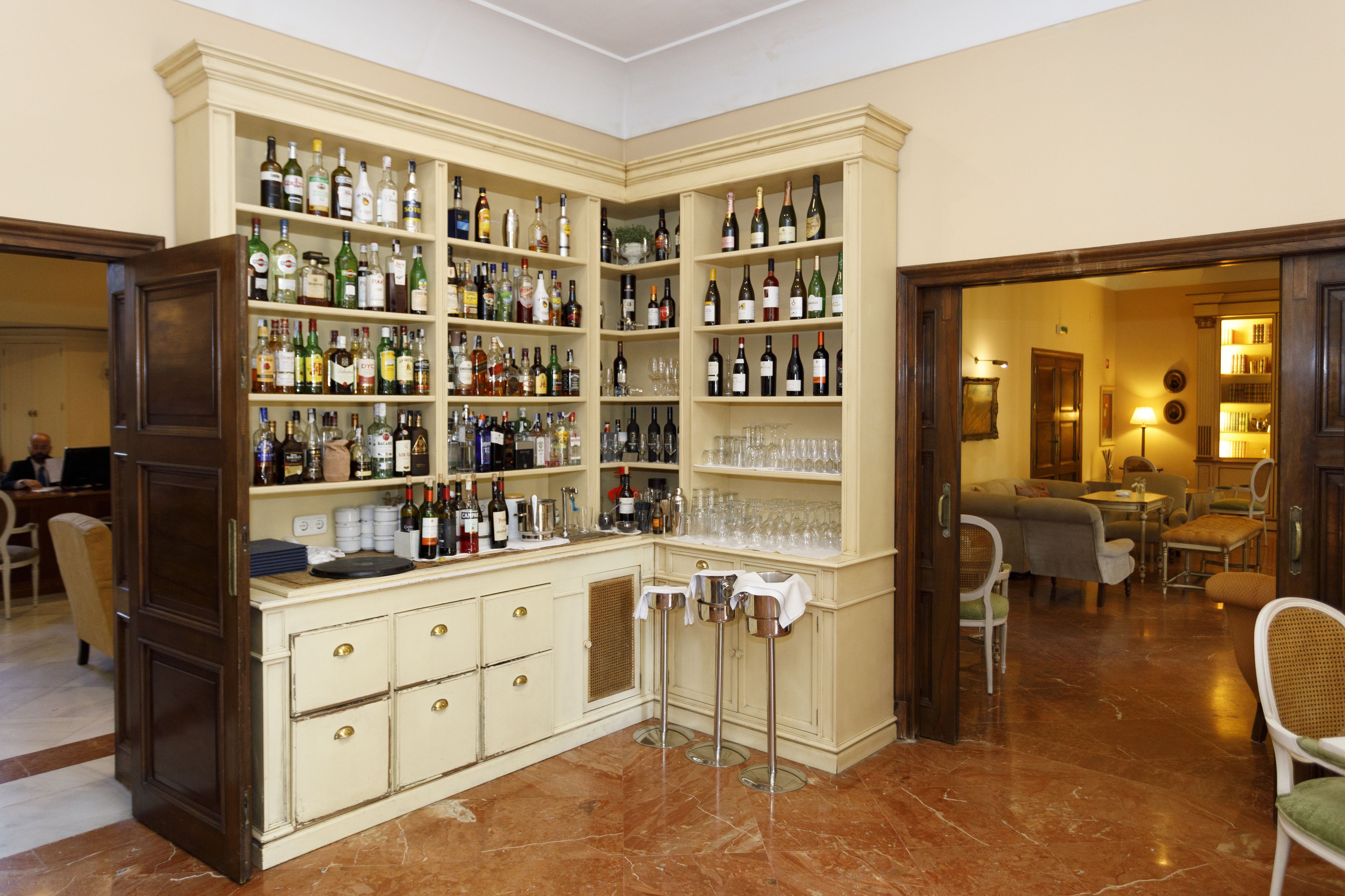 A corner bar area with shelves of liquor bottles and glasses, a bar counter, and seating in the adjacent room.