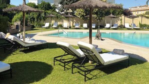 Uma área à beira da piscina com espreguiçadeiras, guarda-sóis e um edifício ao fundo.