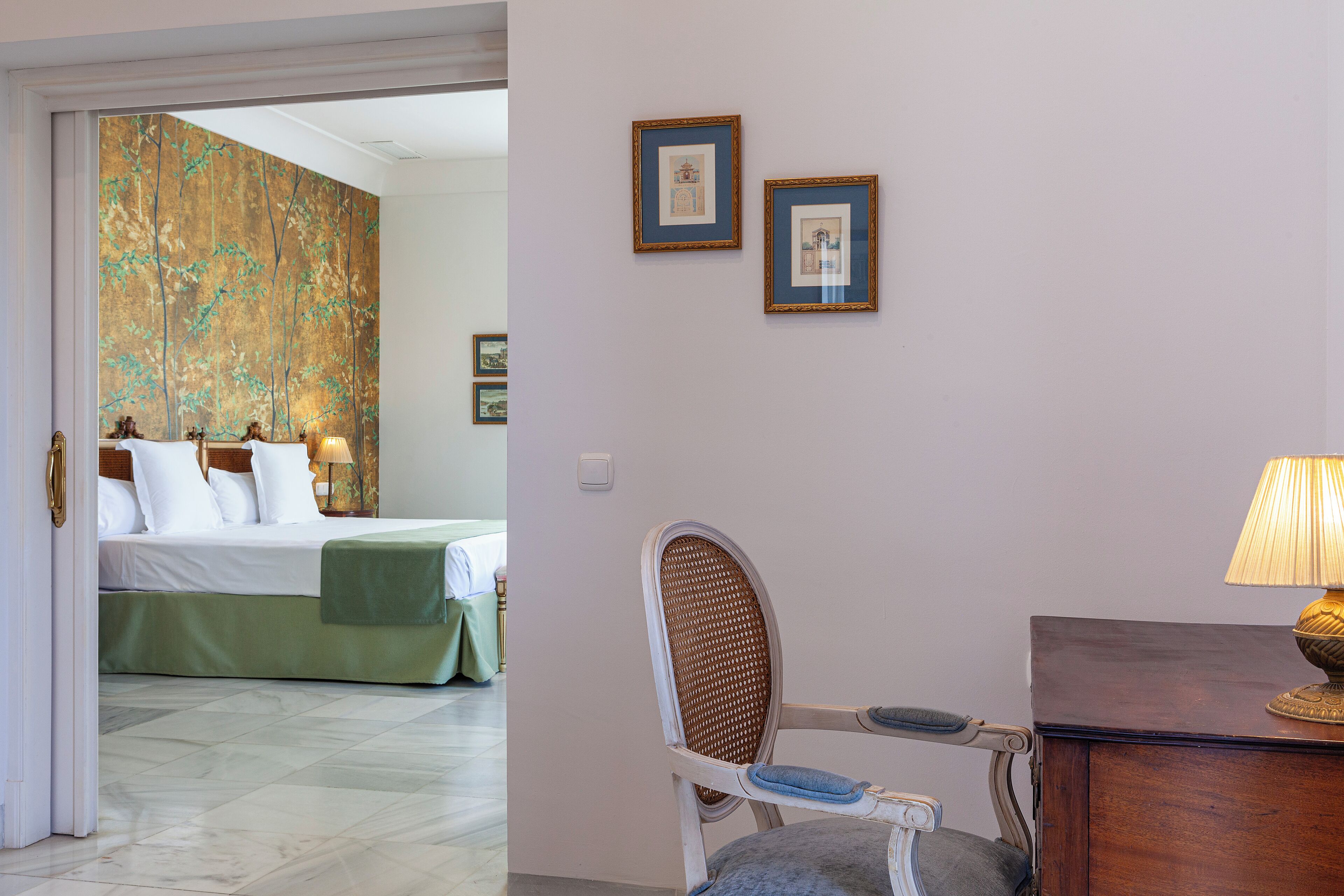 Quarto de hotel com uma cama, uma cadeira, uma escrivaninha e duas fotos emolduradas na parede.