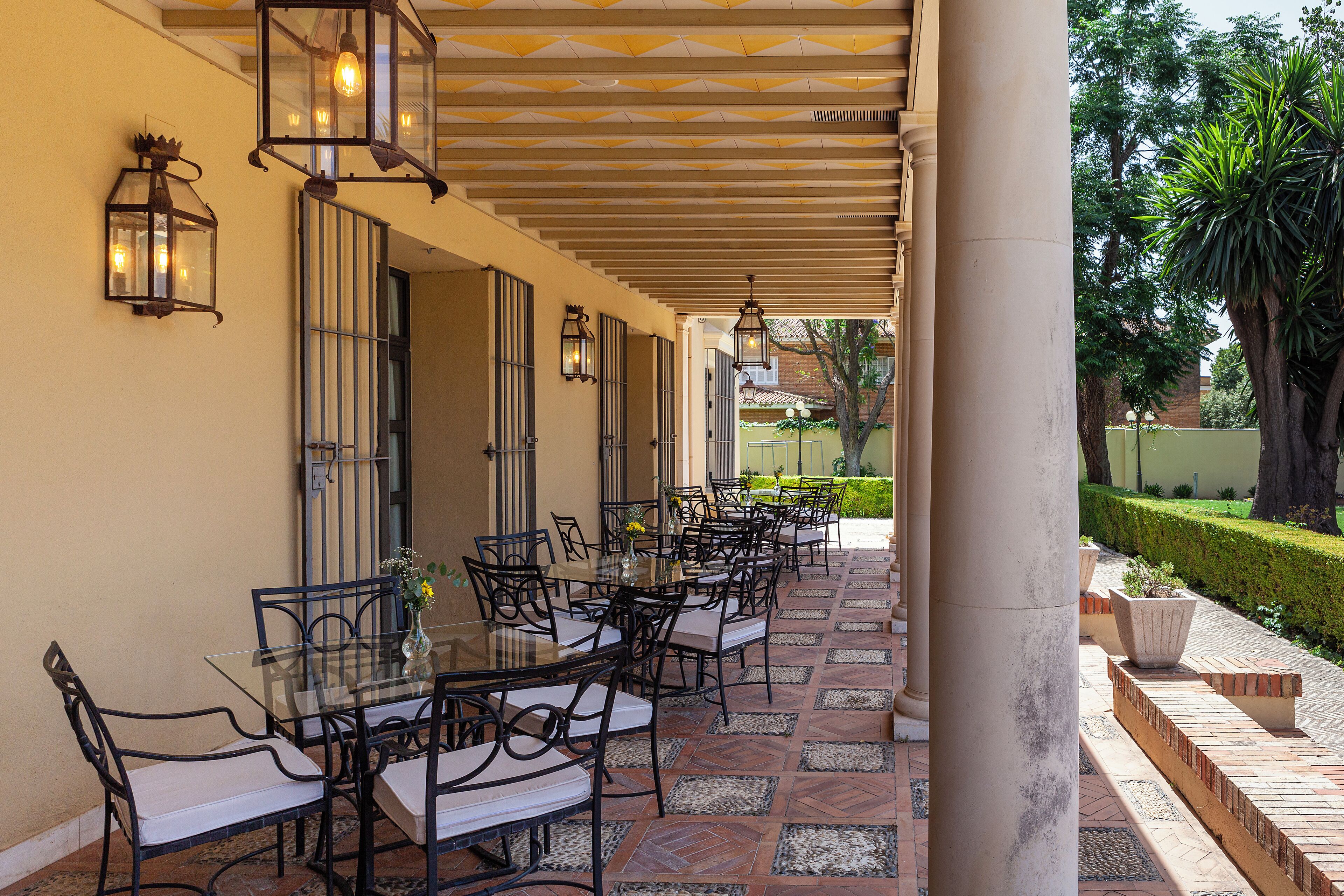 A covered outdoor dining area with glass tables and chairs, surrounded by columns and lanterns.