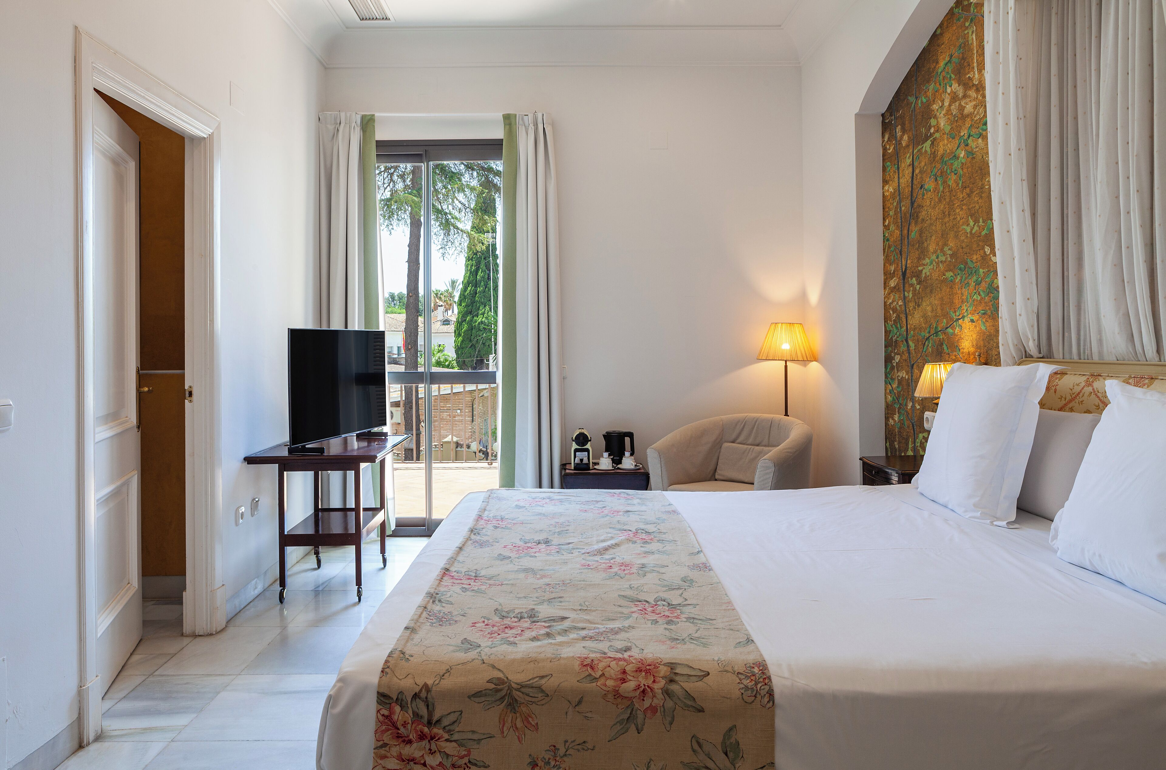 Habitación de hotel con cama, escritorio con televisor, silla, lámpara y vista al exterior.